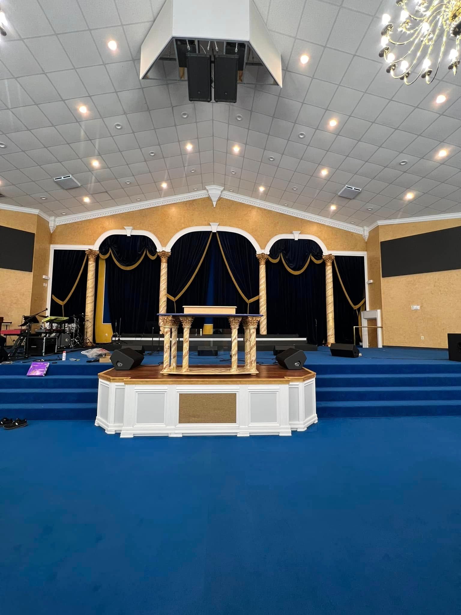 An empty church stage with a raised wooden pulpit, blue carpeting, and navy velvet curtains against gold walls.