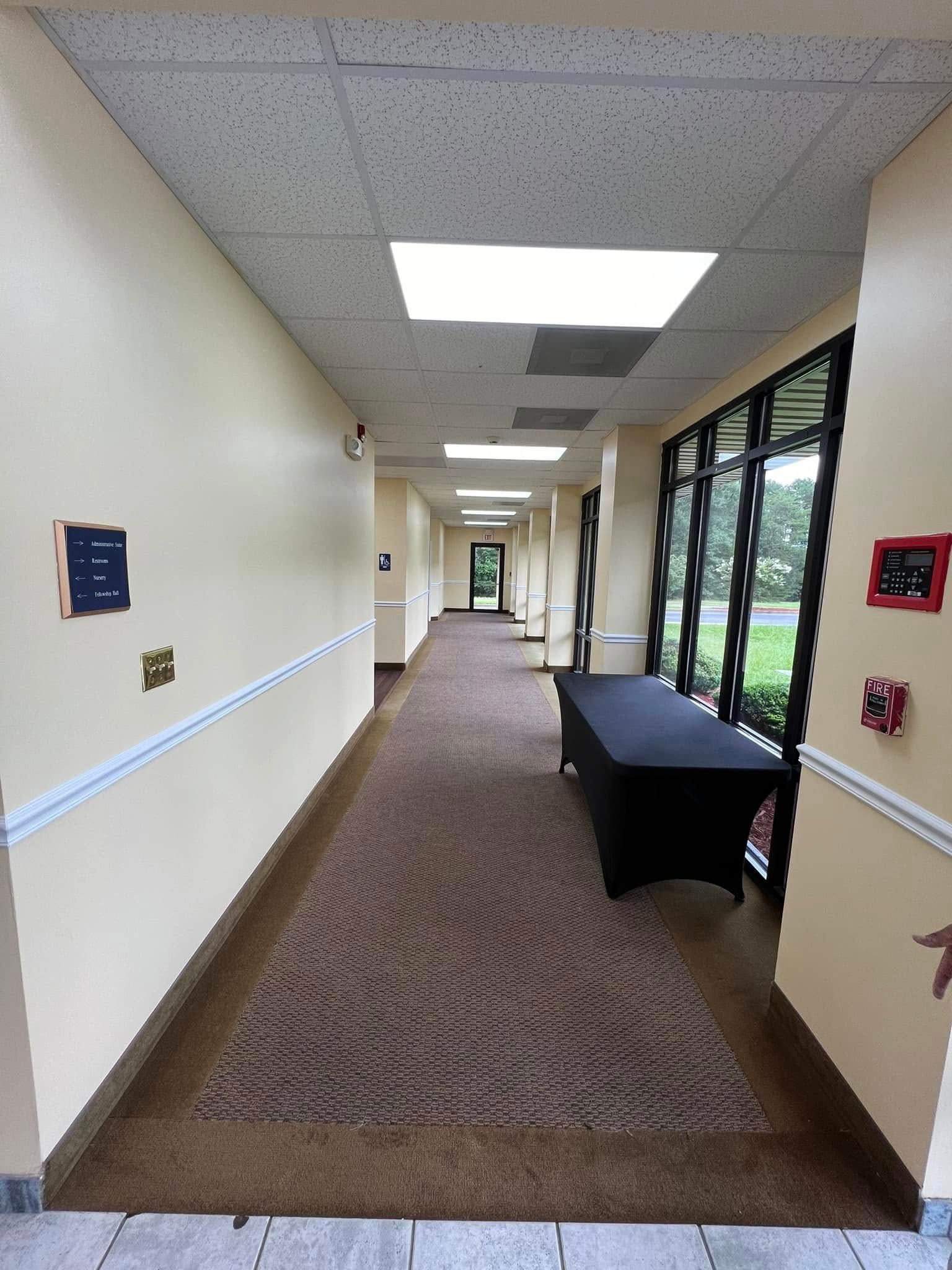 A long, indoor hallway with cream walls, a brown patterned carpet, overhead fluorescent lights, and a black draped table.