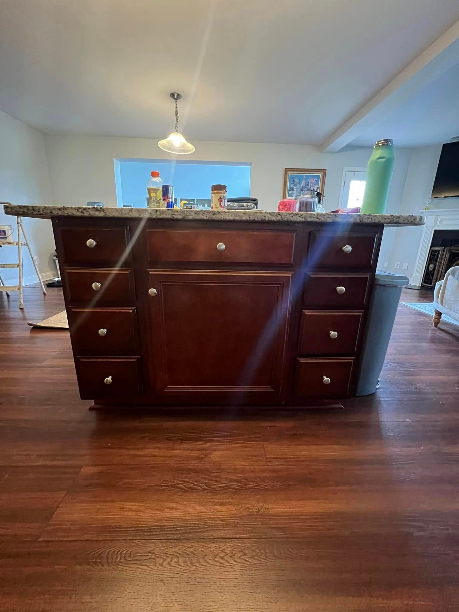 A dark wood kitchen island stands on a wood floor in a home, topped with a granite counter and various items.