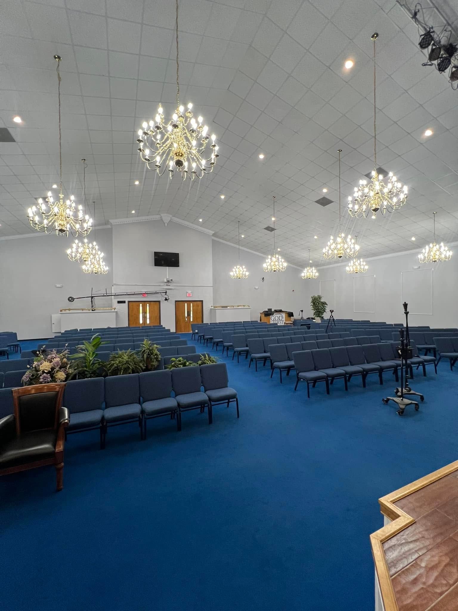 Interior of a church hall with rows of blue chairs, a carpeted floor, and multiple crystal chandeliers on the ceiling.