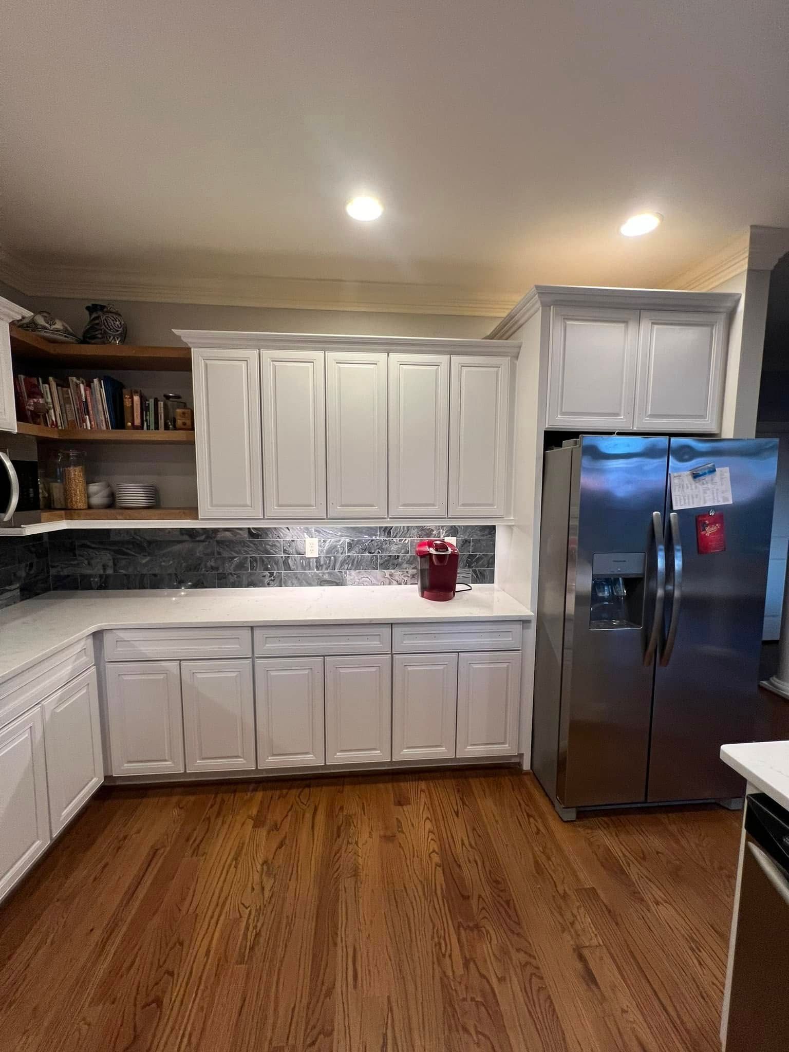 A modern kitchen featuring white cabinets, stainless steel refrigerator, white countertops, and dark hardwood floors.