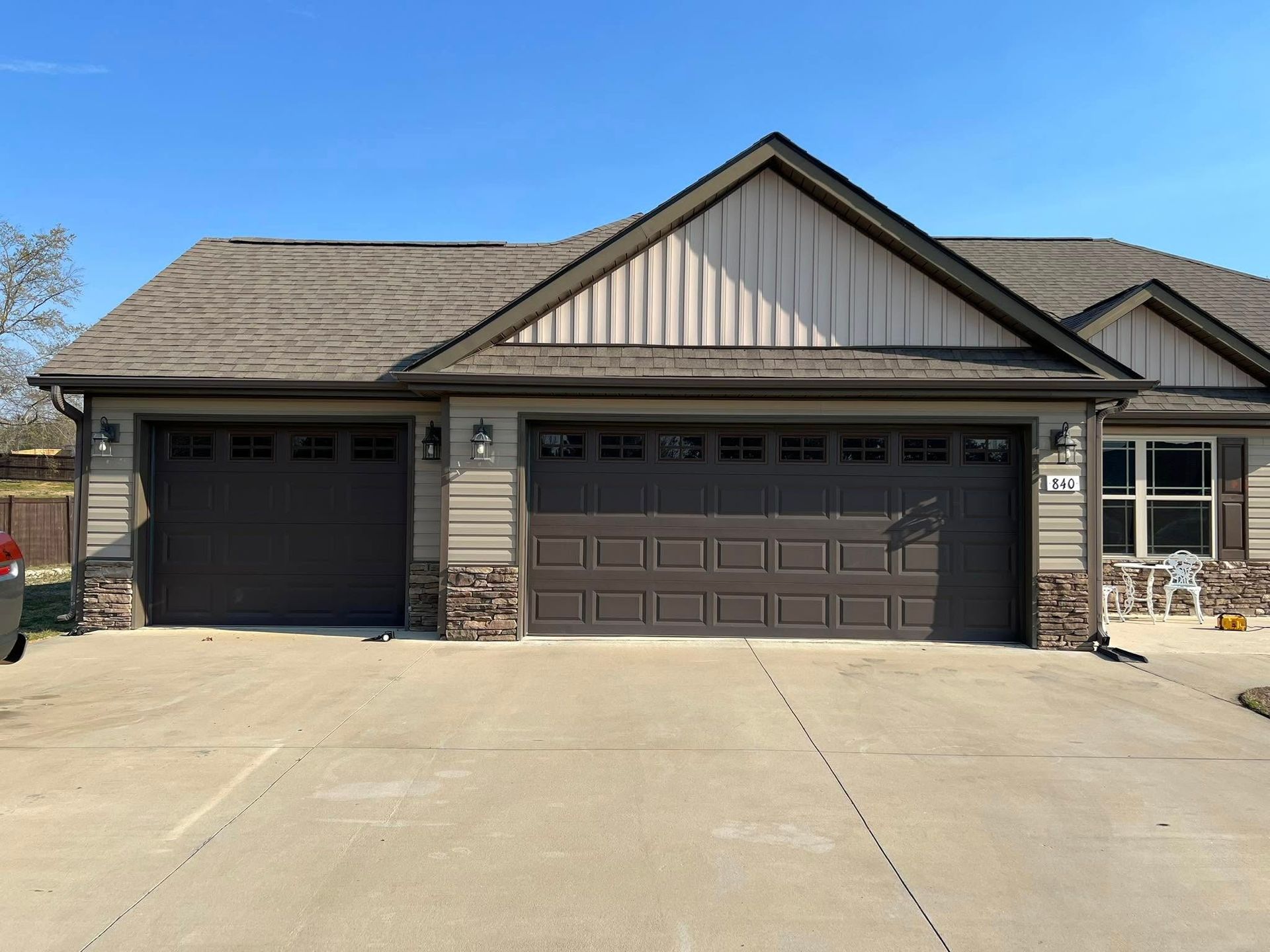 A tan suburban house exterior featuring a two-car garage and a one-car garage, both with brown doors, under a blue sky.