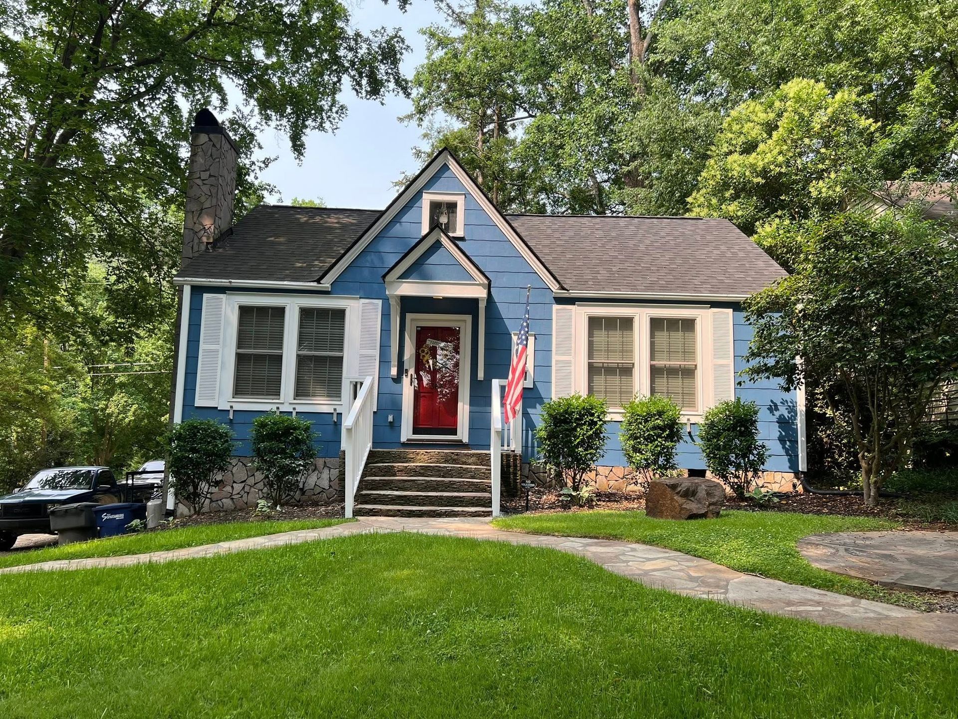 A blue, one-story house with a red front door, white trim, a stone foundation, and a stone walkway in a tree-filled yard.