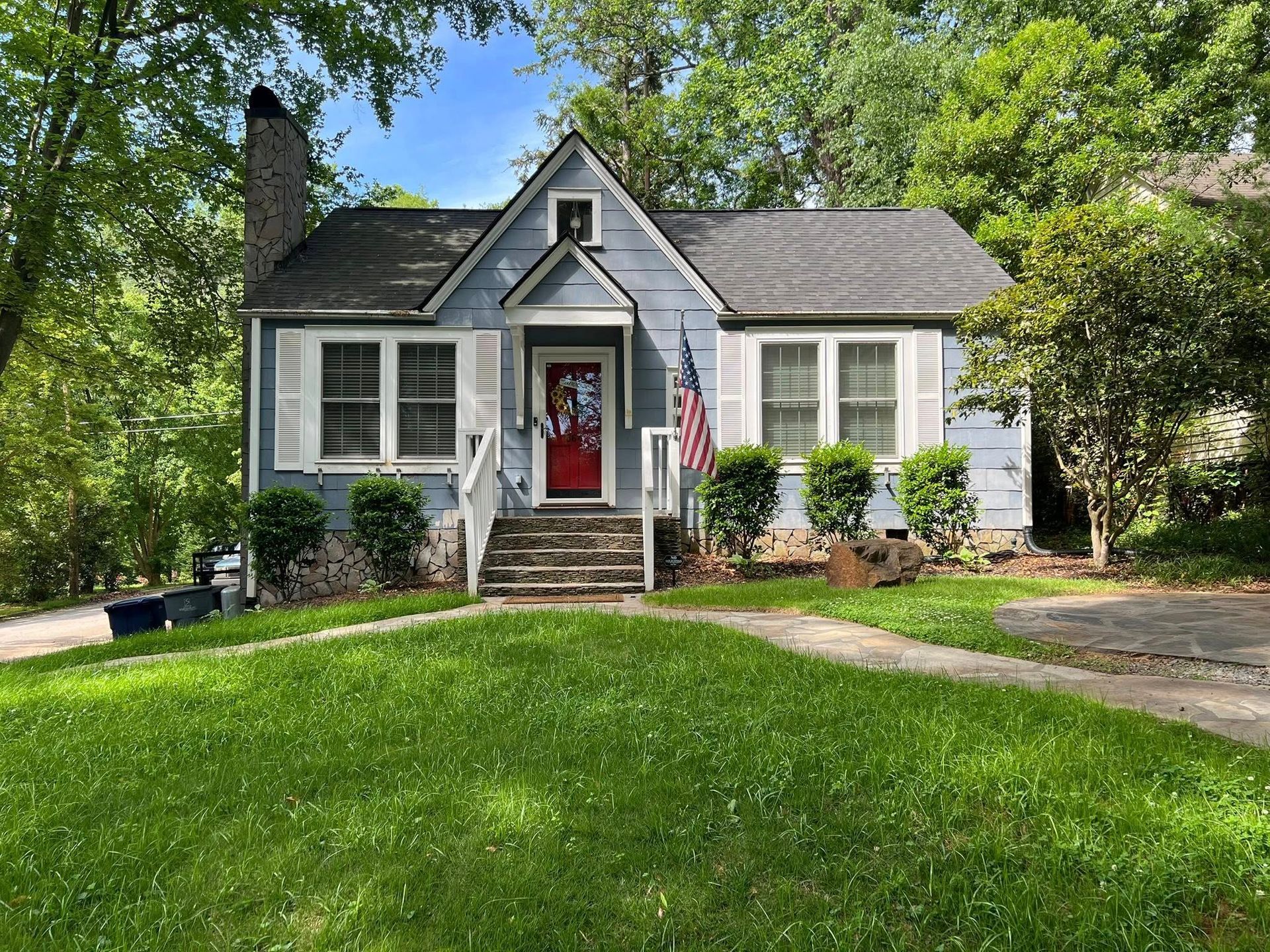 A single-story blue cottage with white trim, a red front door, stone steps, and a grassy front lawn under sunny skies.