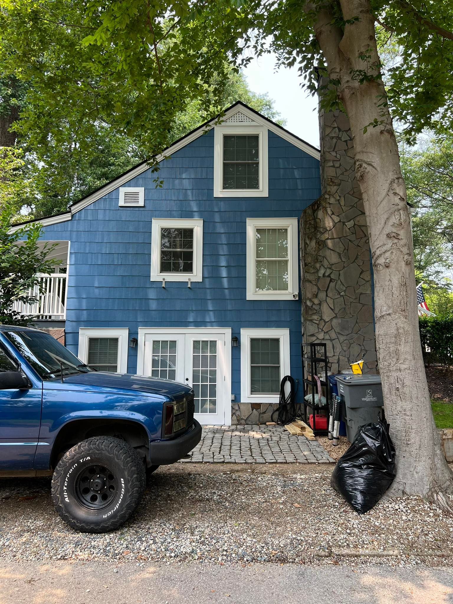 A blue house with white trim stands beside a large tree, with a blue truck parked in the gravel driveway in front.