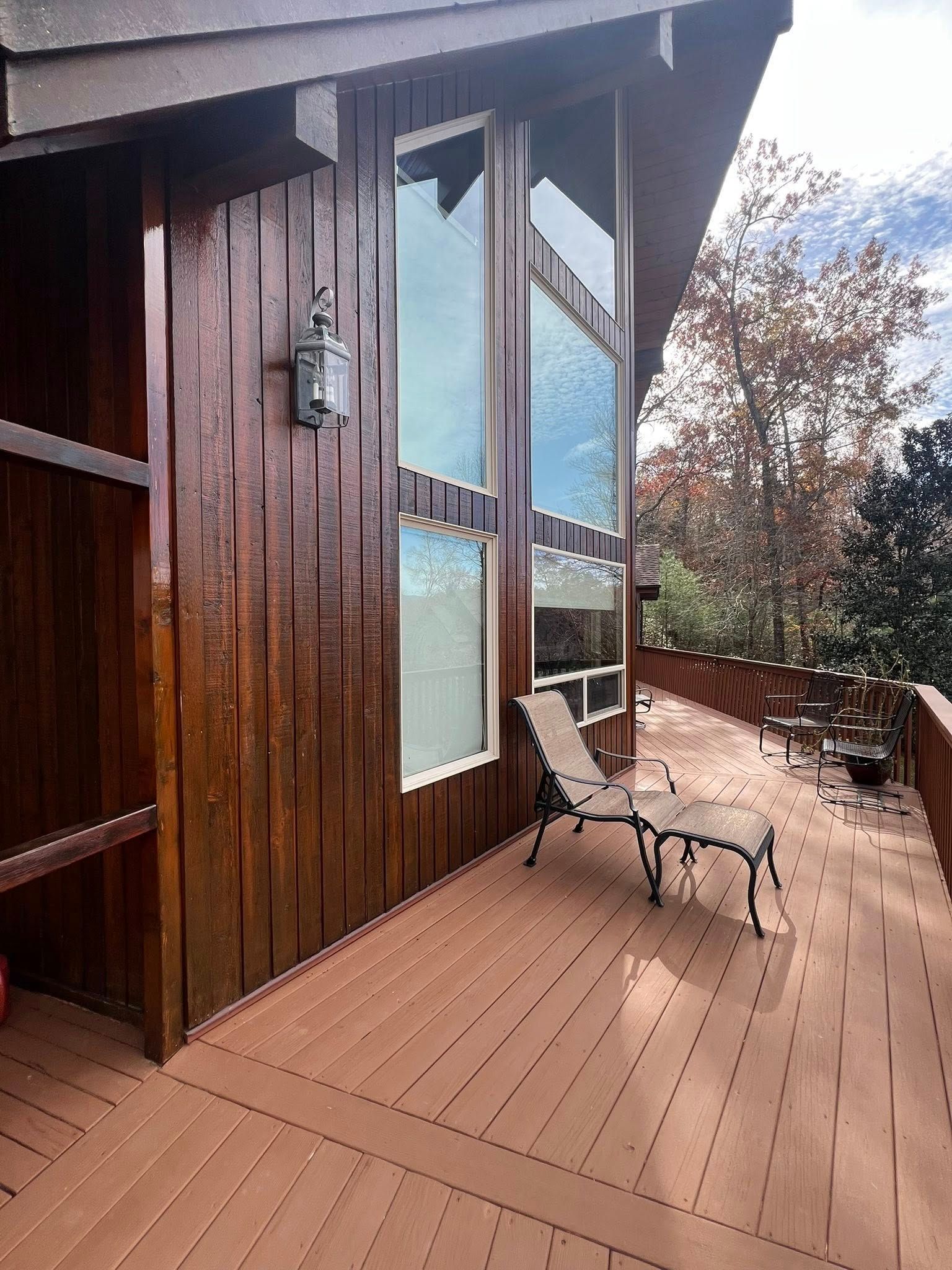A lounge chair sits on a brown wood deck beside a rustic cabin wall with tall windows and a wall-mounted outdoor light.