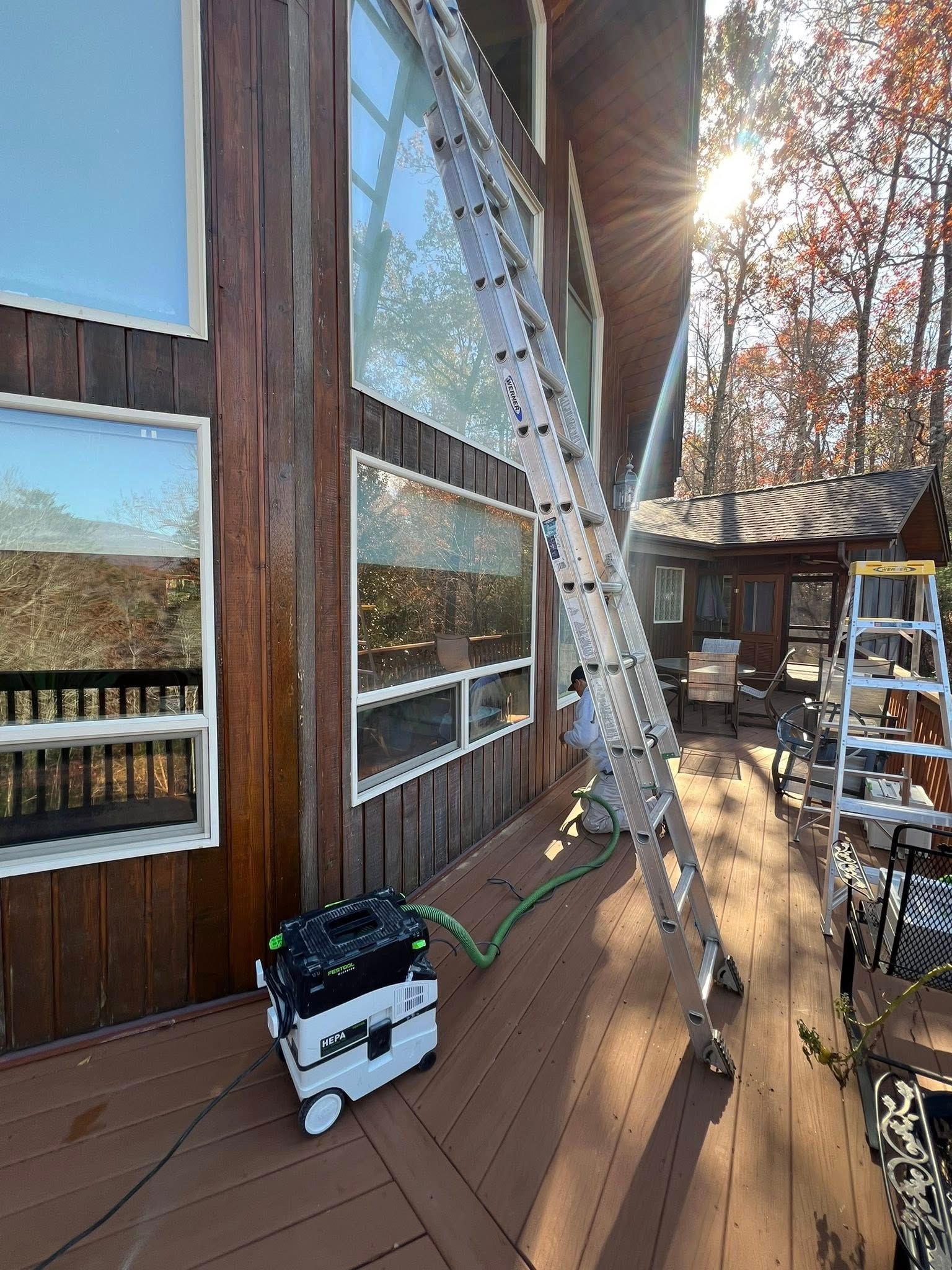 A tall ladder leans against a wood-paneled home, connected to a Festool vacuum on a deck near outdoor furniture.
