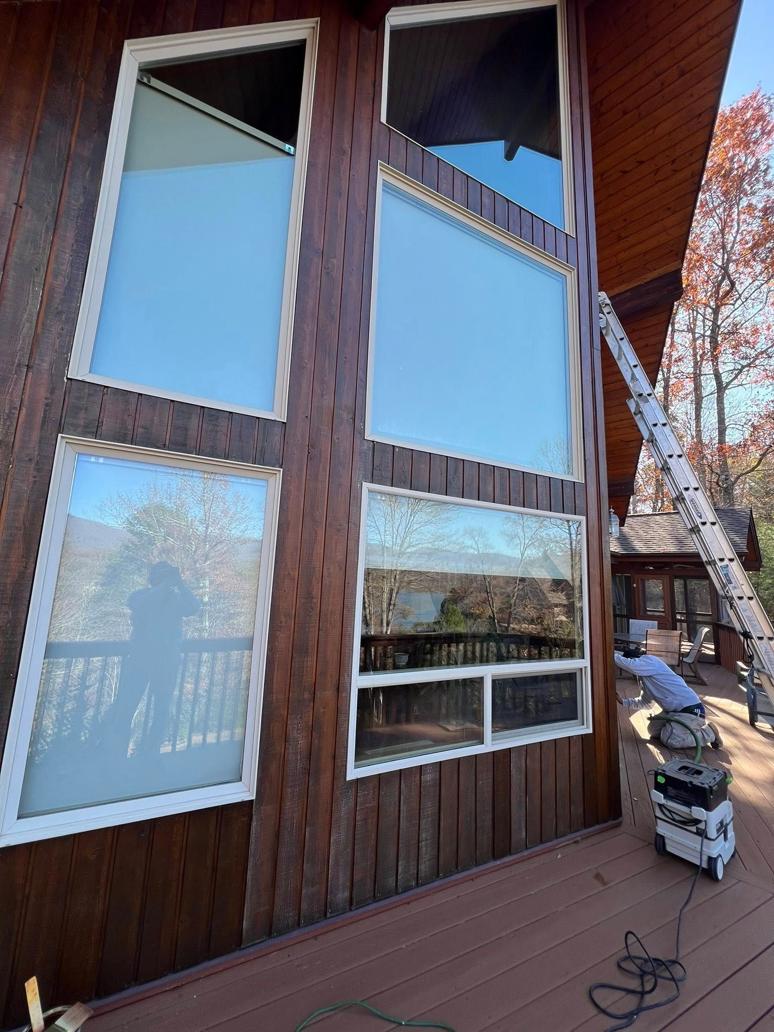 A ladder leans against the exterior of a brown wood-paneled house with large, floor-to-ceiling windows on a deck.