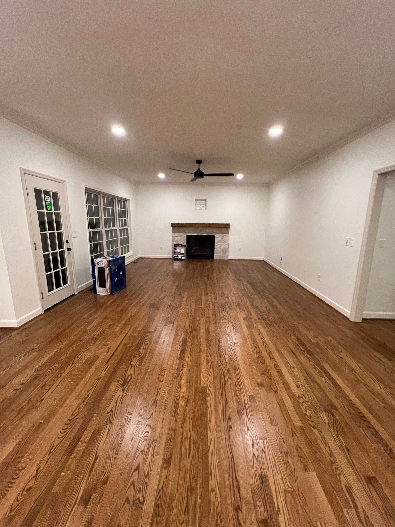 A spacious, empty living room with polished hardwood floors, white walls, recessed lighting, and a brick fireplace.