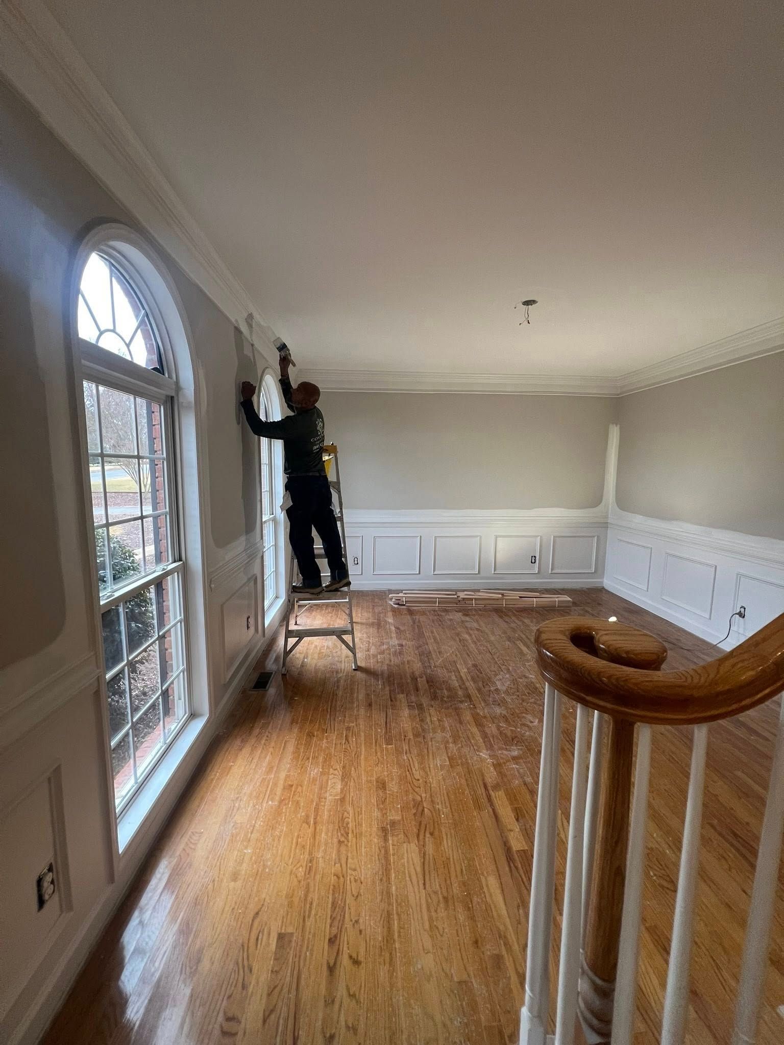 A person stands on a ladder, painting the wall near a large arched window in a room with hardwood floors and wainscoting.