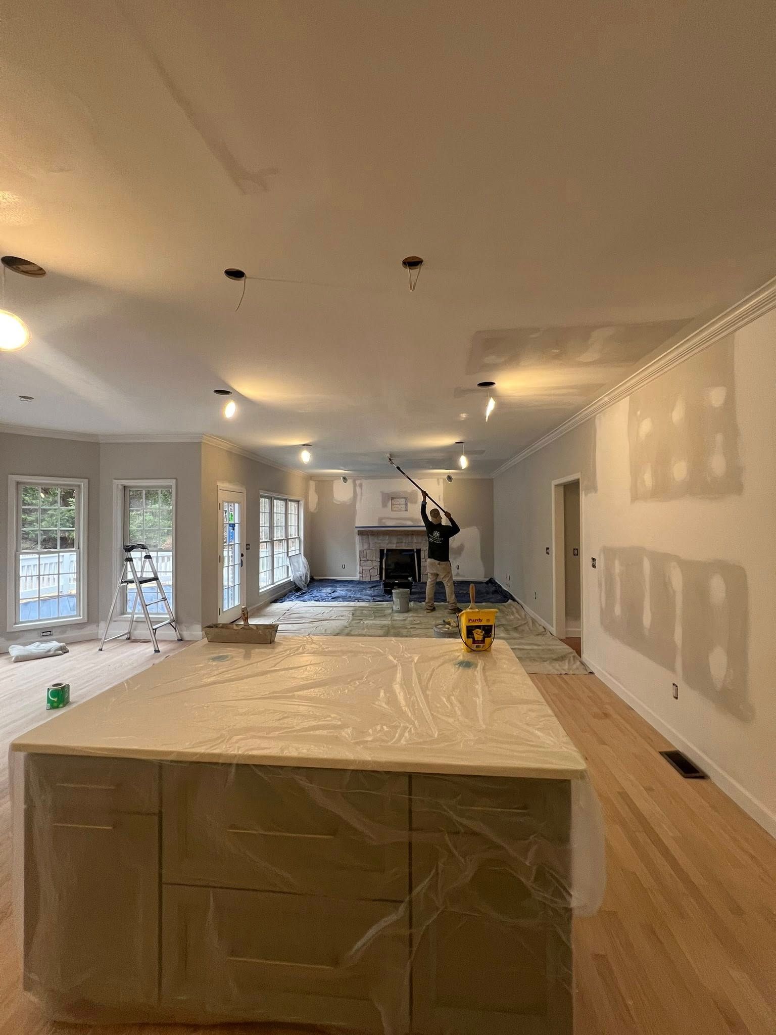 A person stands in a large, unfinished room with hardwood floors, using a long-handled tool to sand a white ceiling.