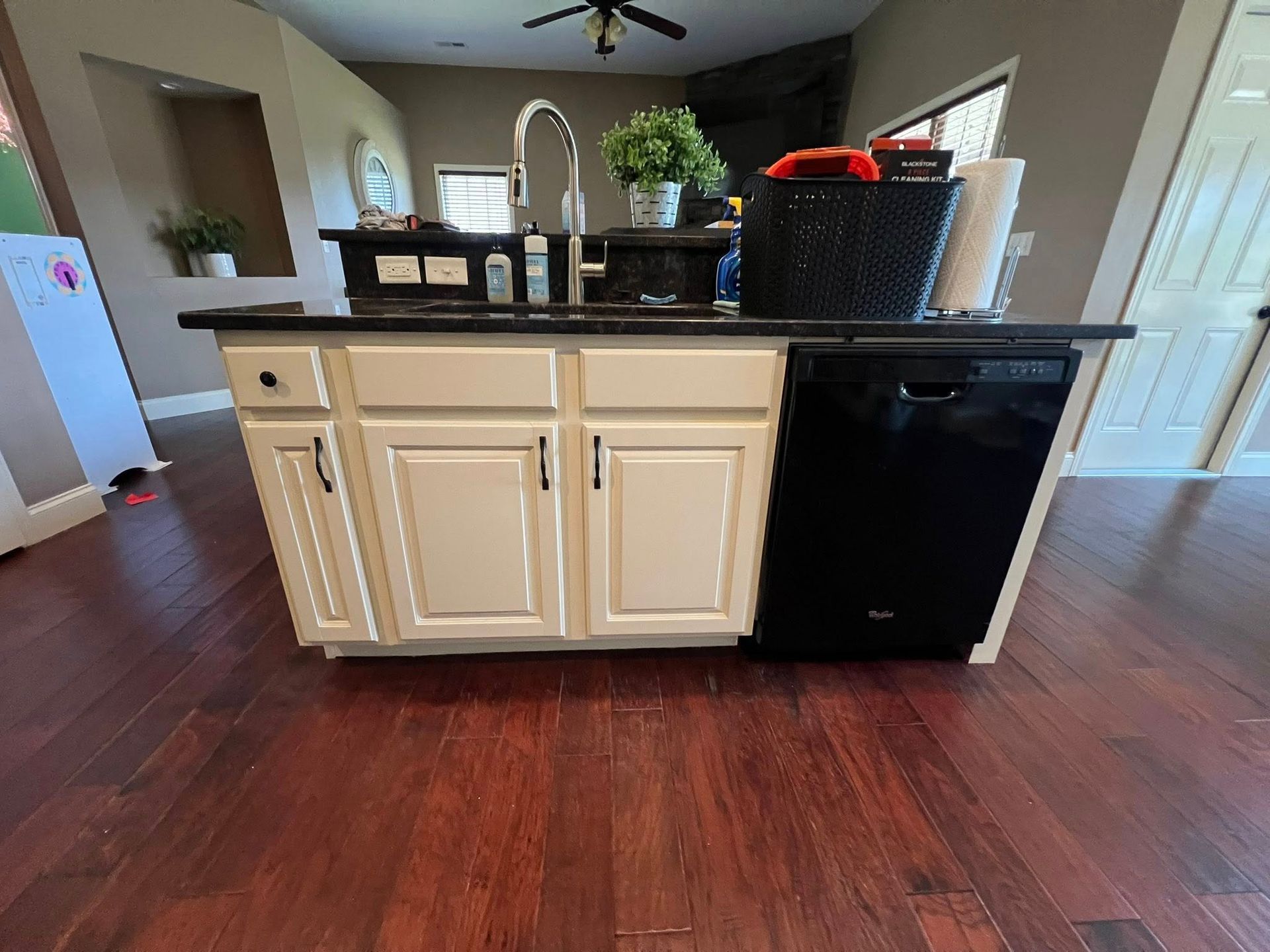 A kitchen island with white cabinets, a black countertop, and a black dishwasher set on dark wood flooring.