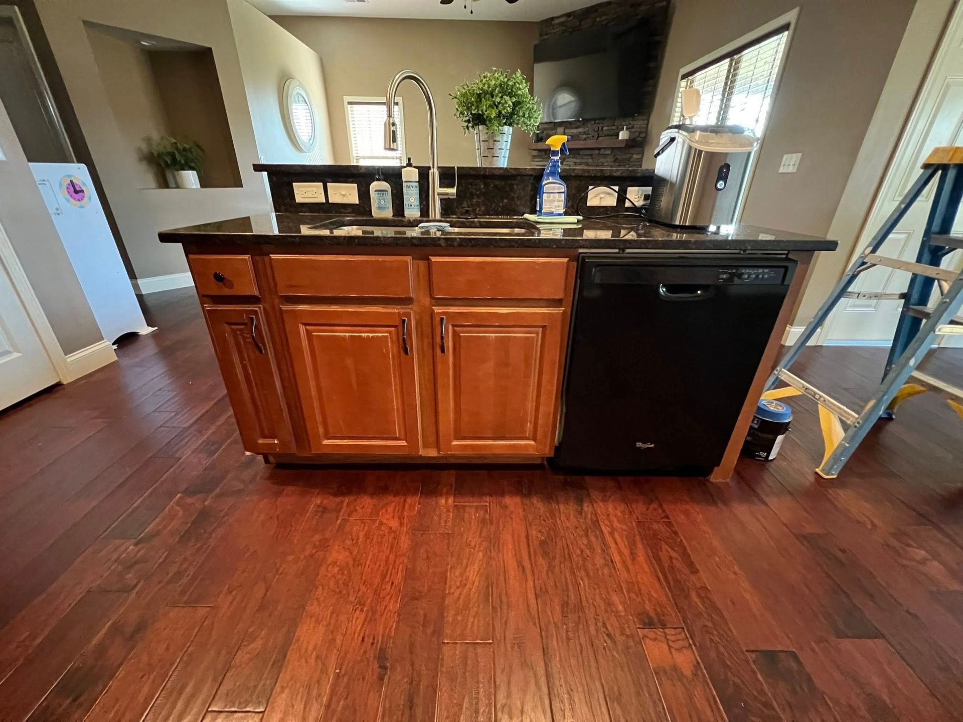 A kitchen island with brown wooden cabinets, a black dishwasher, a dark countertop, and a faucet on hardwood flooring.