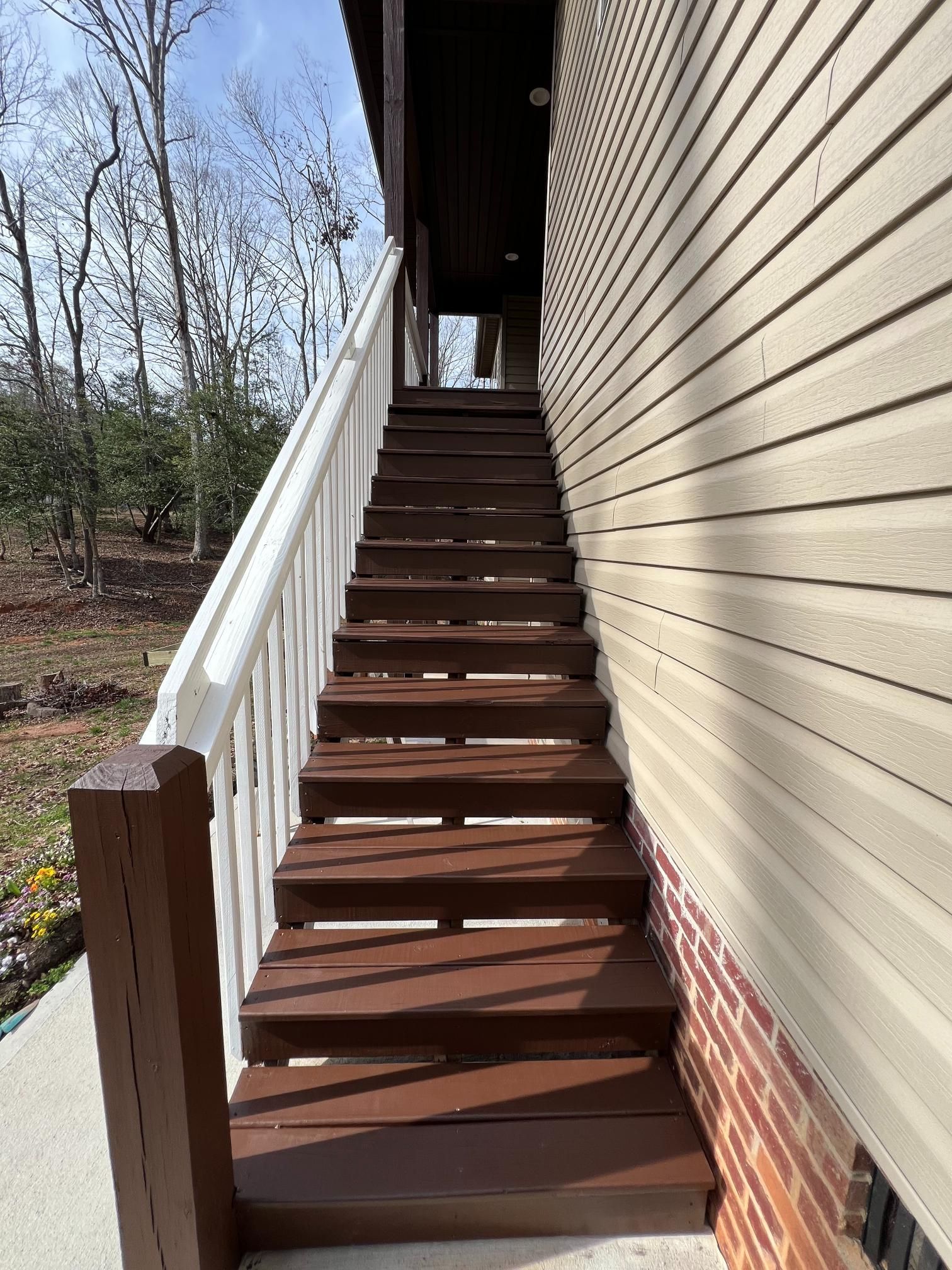 A set of brown wooden stairs with a white railing leading up to an elevated deck on the side of a house.