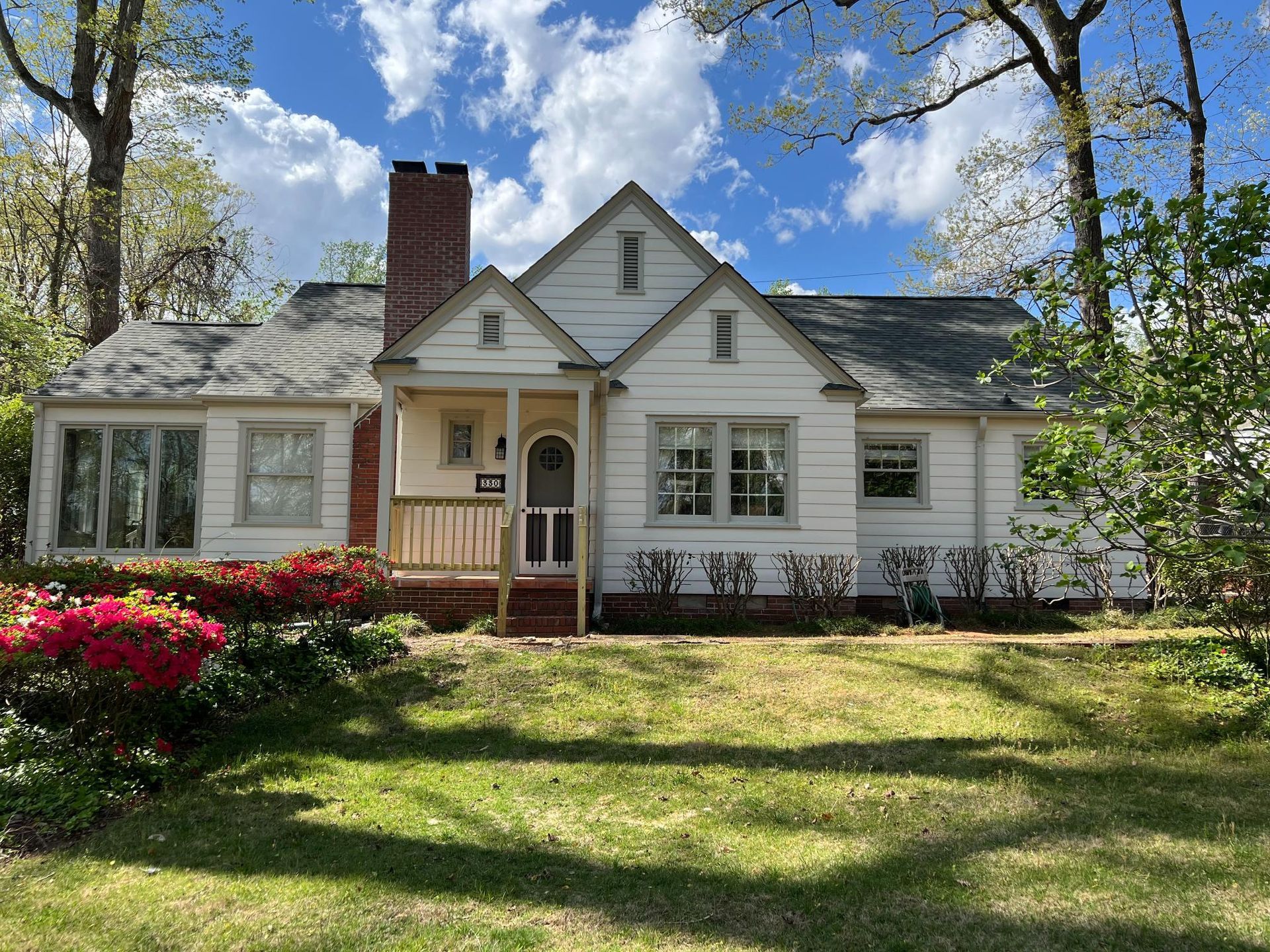 A white single-story cottage with a brick chimney and a small front porch, surrounded by green trees and red flowers.