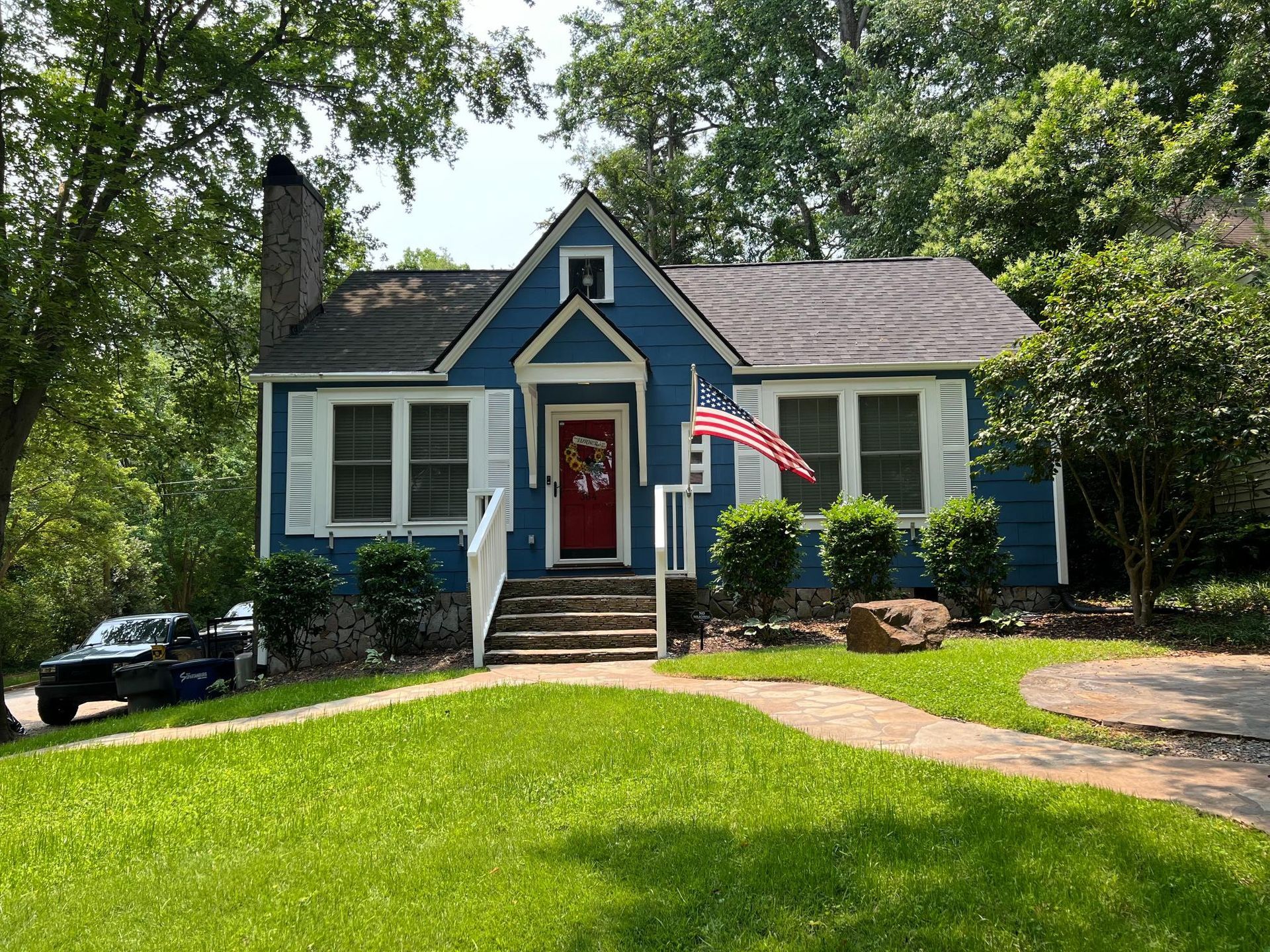 A blue cottage-style house with a red front door, white trim, and an American flag, set behind a grassy lawn.