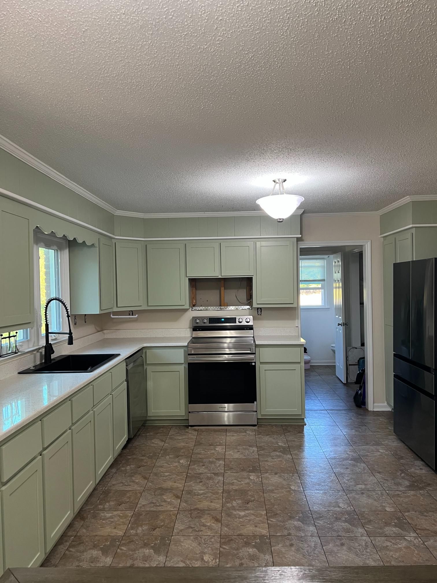 A kitchen with light sage green cabinets, black appliances, a white countertop, and tan tile flooring.