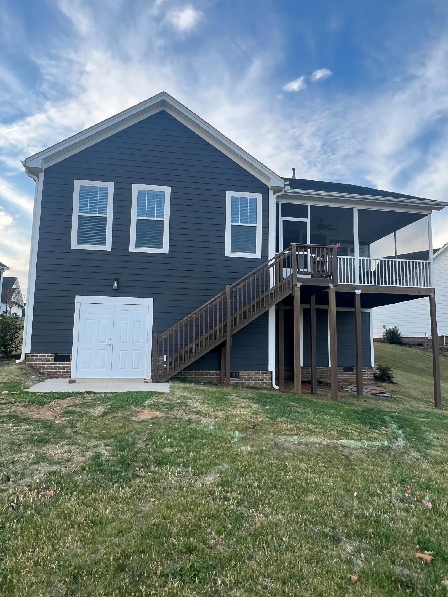 A two-story dark blue house with a white lower-level door, an elevated screened porch, and wooden stairs on a grassy hill.