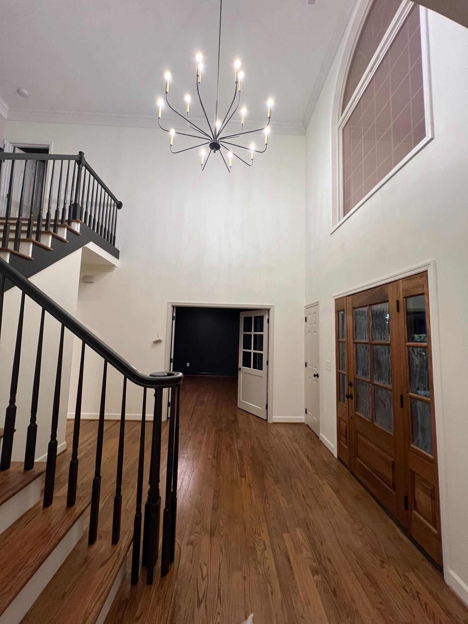 A grand, high-ceilinged foyer with hardwood floors, a staircase with black railings, a modern chandelier, and wood doors.