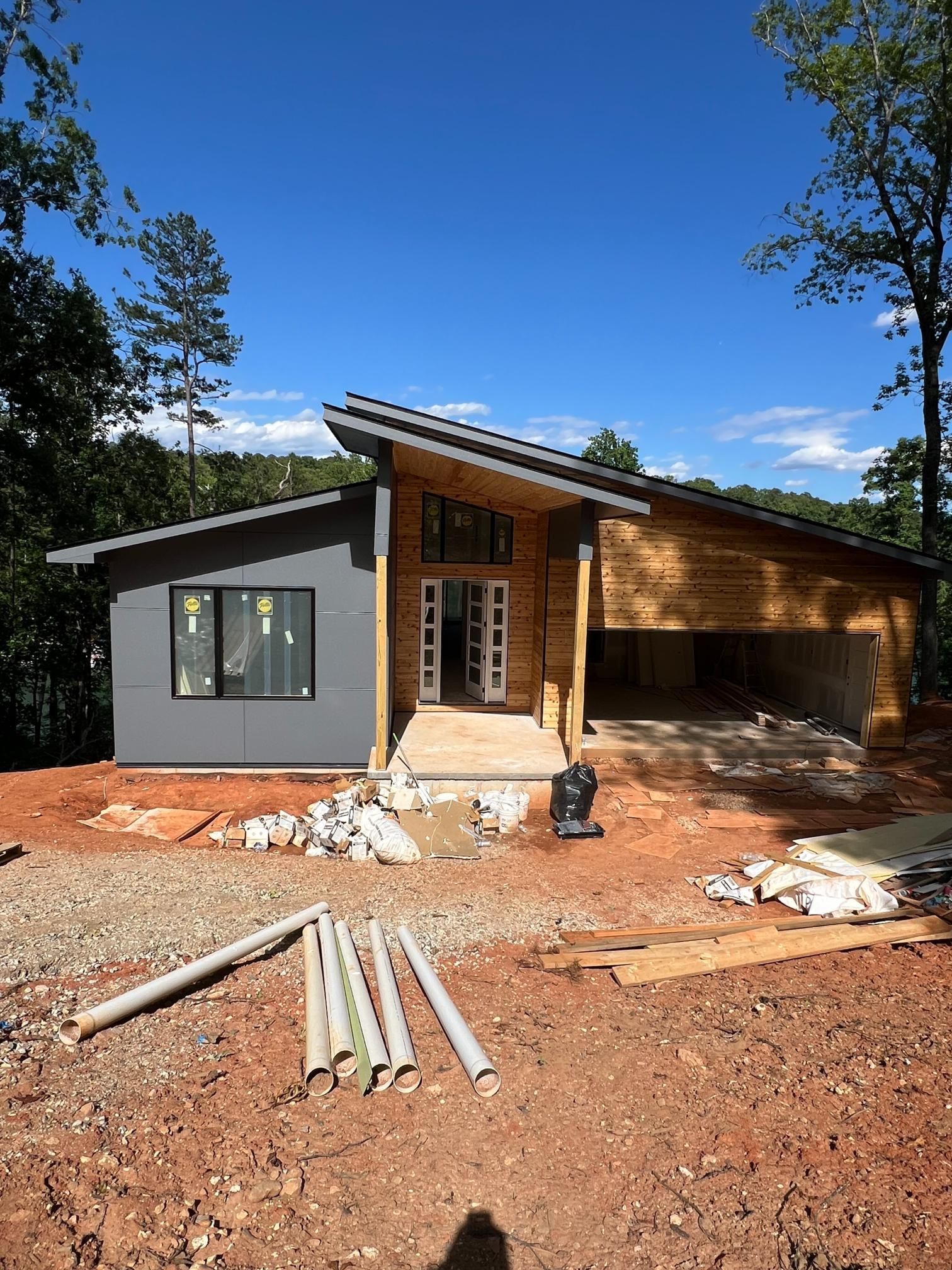 A modern house under construction with a grey exterior, wood accents, and a slanted roof on a dirt lot with blue sky.