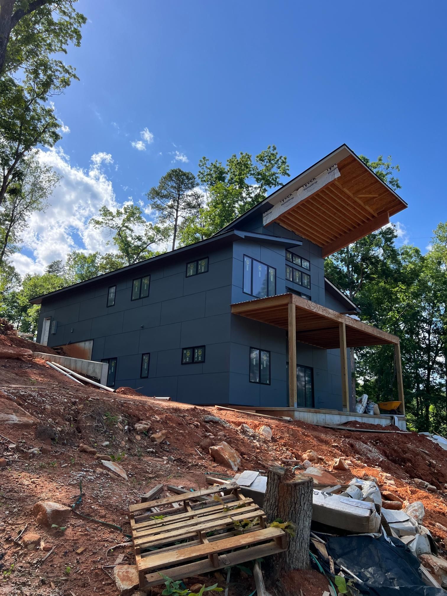 A two-story, charcoal-colored house with a cantilevered roof and wooden deck stands on a red dirt hillside among trees.