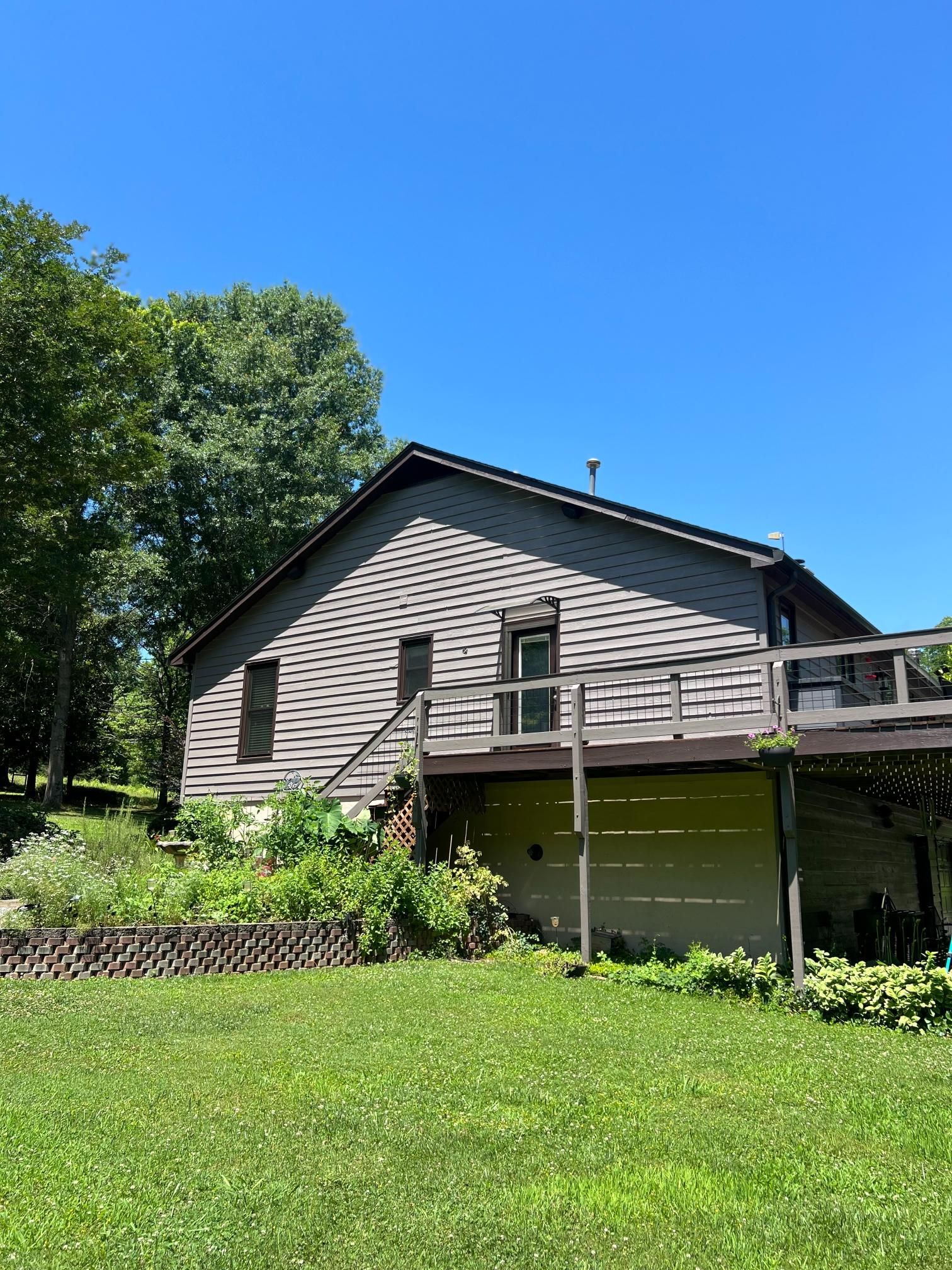 A gray-sided house with a large wooden deck stands next to a lush green yard under a clear blue sky.