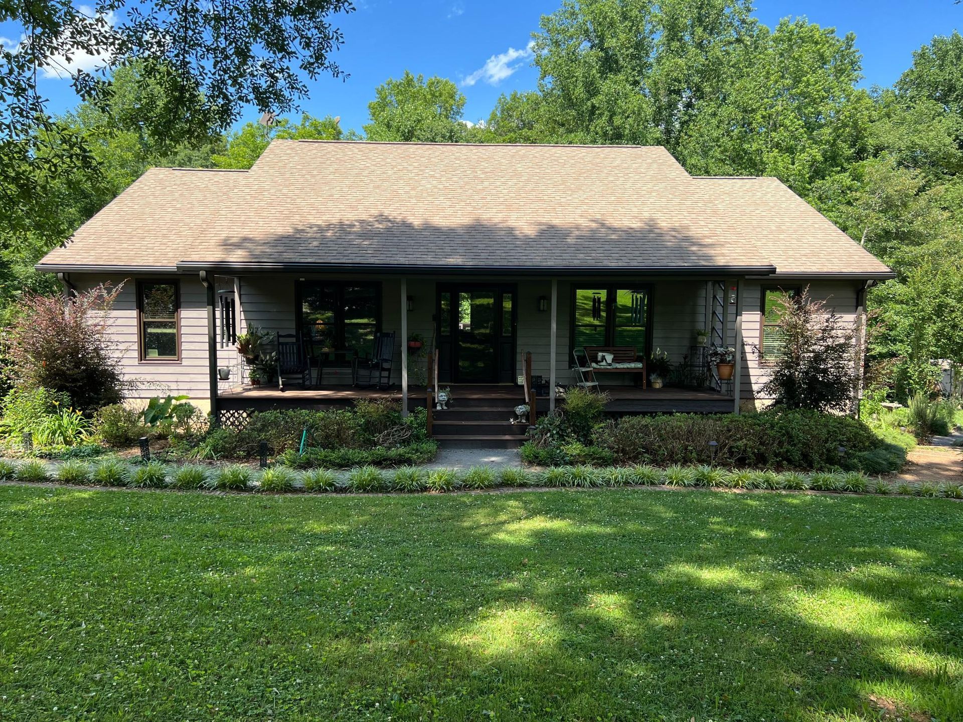 A tan-sided house with a large front porch and a dark shingled roof, surrounded by lush green trees and a lawn.