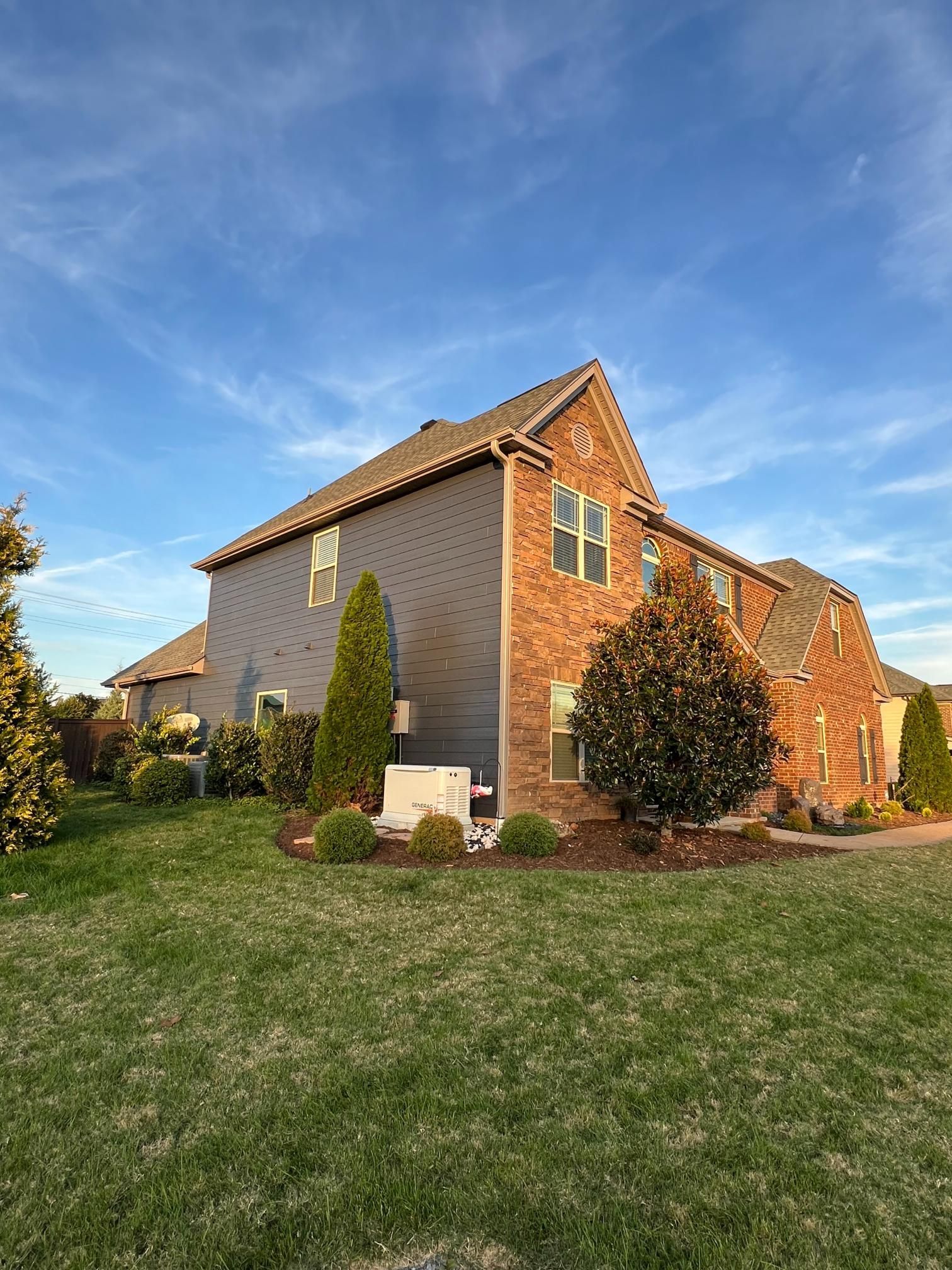 A side view of a brick house with dark gray siding, green landscaping, and a backup generator under a blue sky.