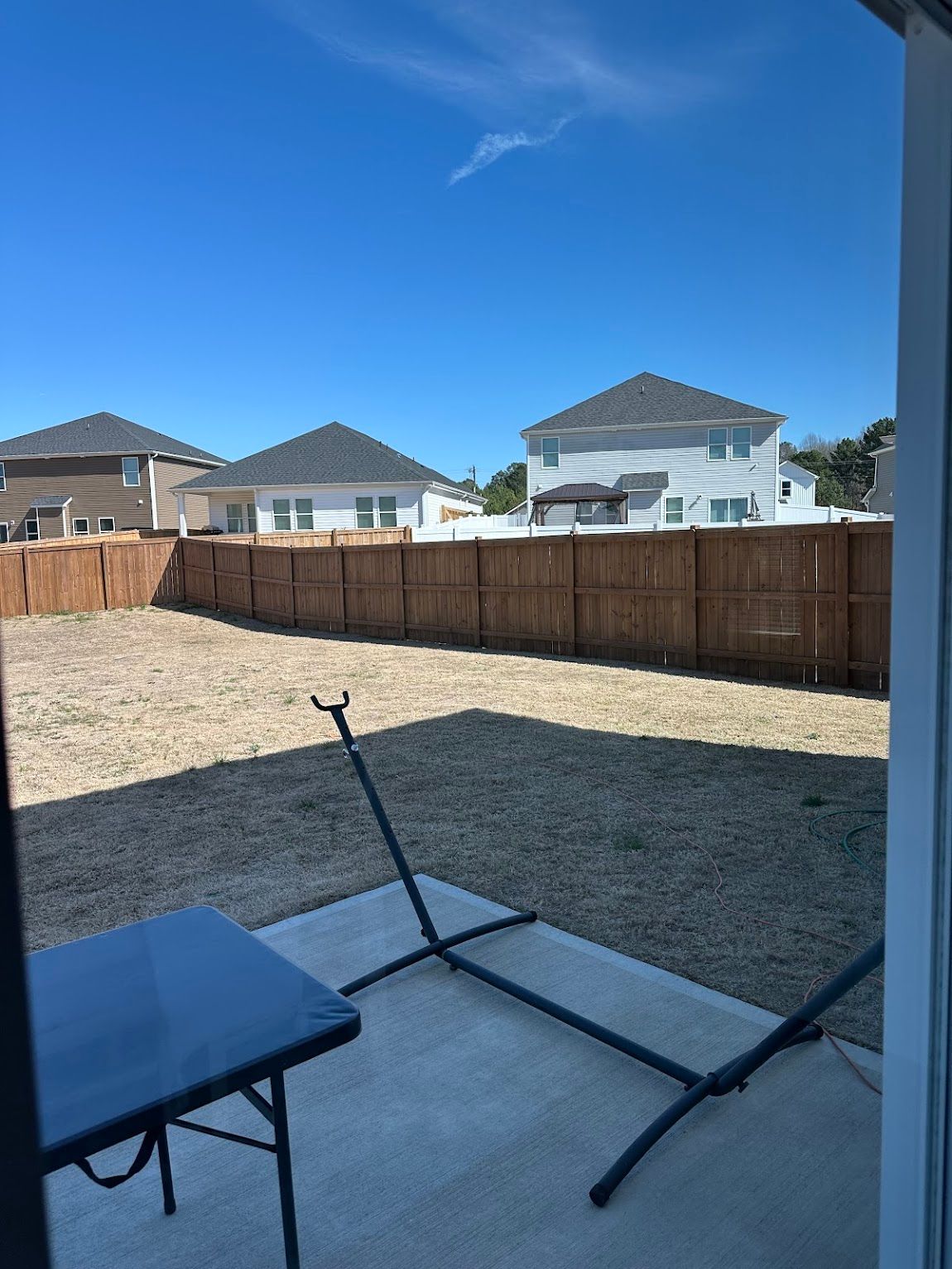 A backyard with a stone patio, a small table, a hammock frame, and a wooden fence under a clear blue sky.