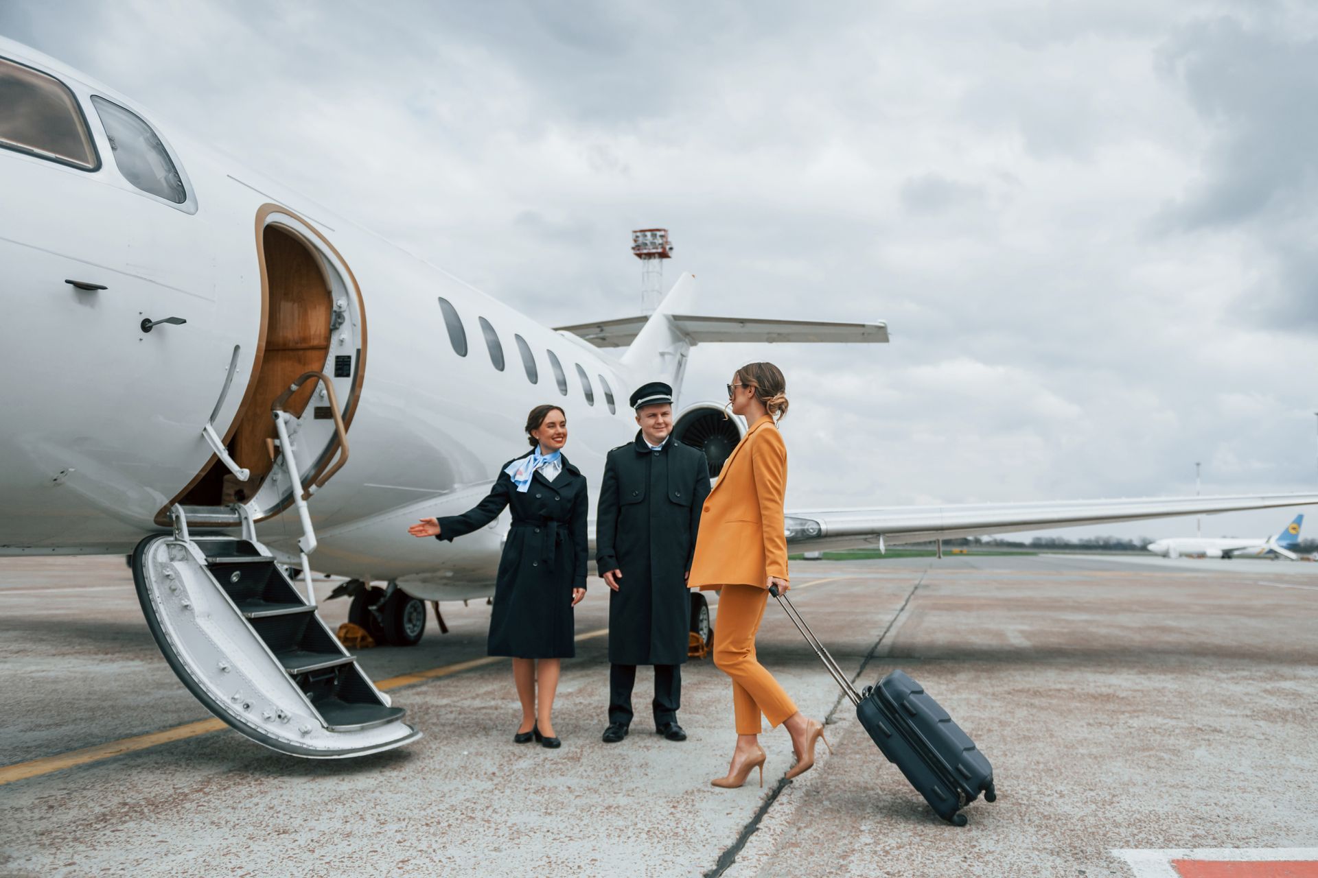 Woman in orange suit boards a private jet with luggage, greeted by pilot and flight attendant.