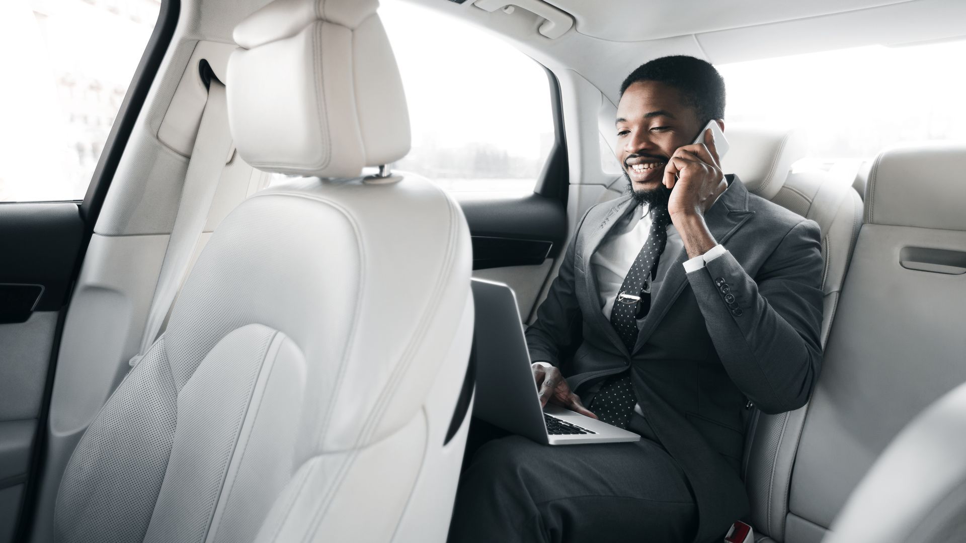 A man in a suit works on a laptop and talks on the phone in the backseat of a car.