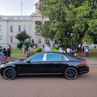 Black luxury car parked in front of a courthouse, people gathered nearby.