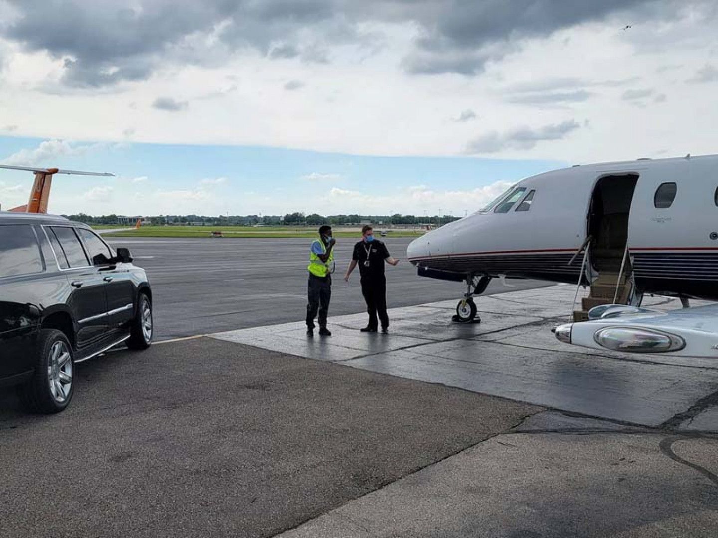 Two airport workers beside a private jet on a tarmac. A black SUV is parked nearby.