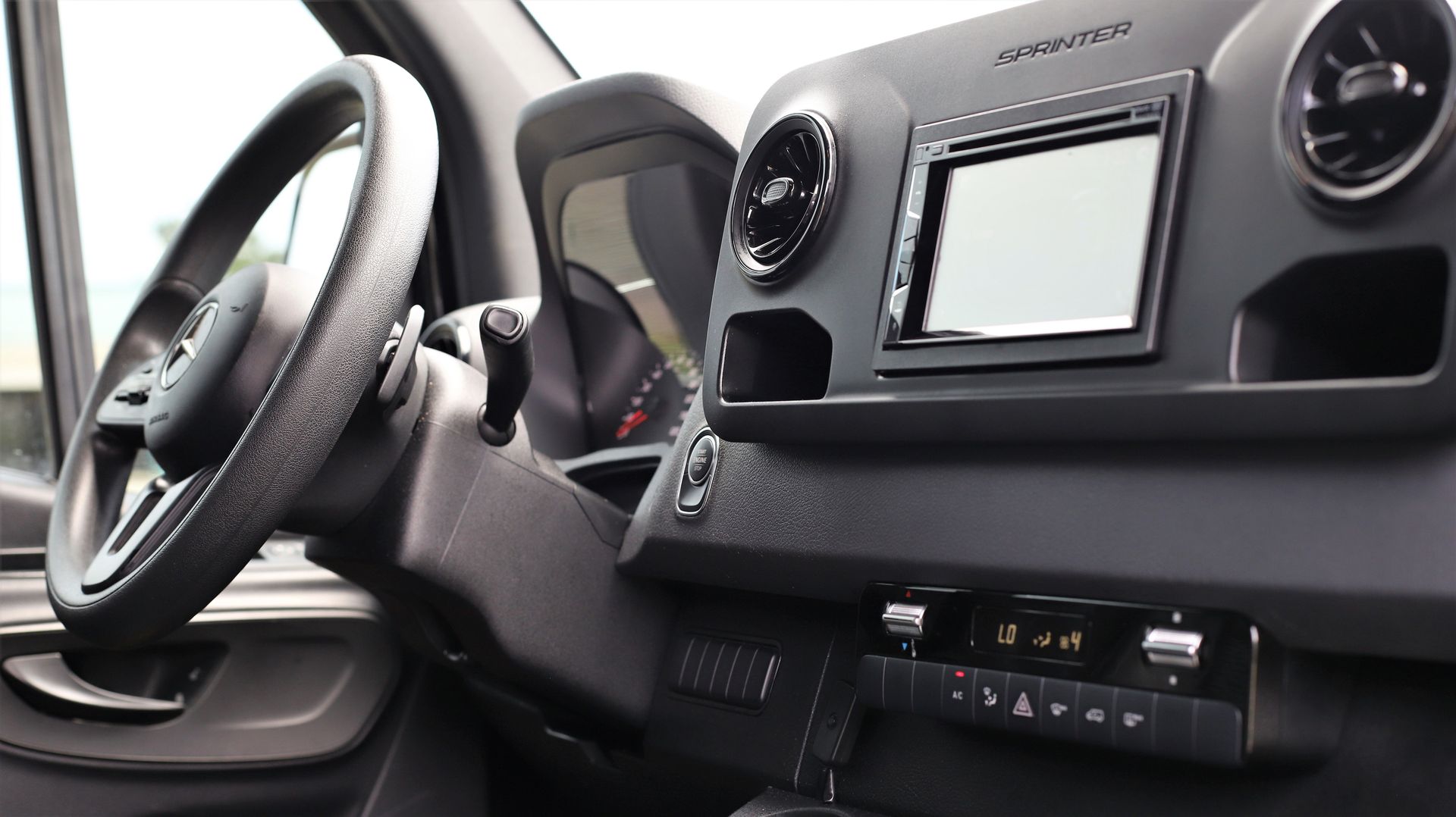 Interior view of a vehicle dashboard with steering wheel, screen, and climate controls. Black and gray colors.