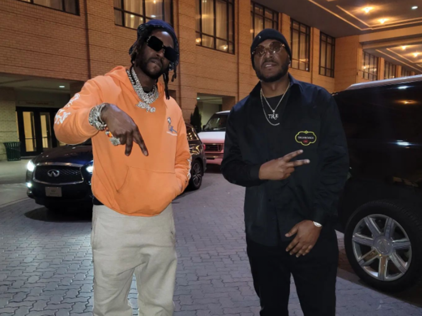 Two men posing outside a building; one in orange hoodie, the other in black jacket, making hand gestures.