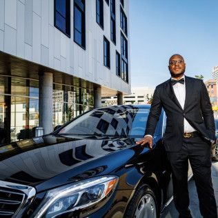 Man in a suit stands next to a black car in front of a modern building.