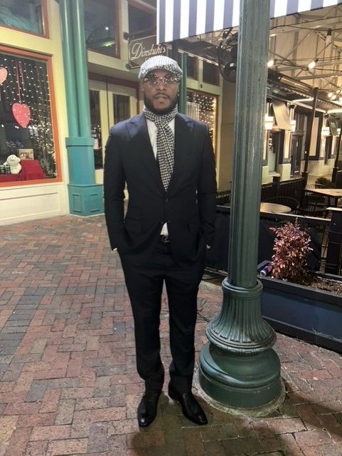 Man in a suit and cap stands near a lamp post. Brick patio, restaurant in the background.