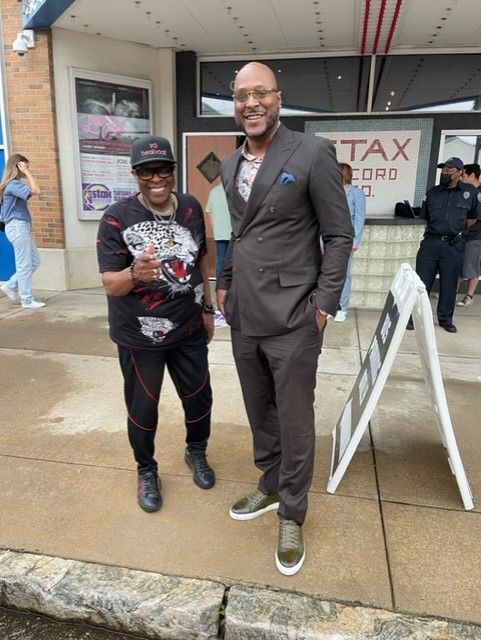 Two men pose outside a building. One wears a suit, the other a t-shirt and track pants.