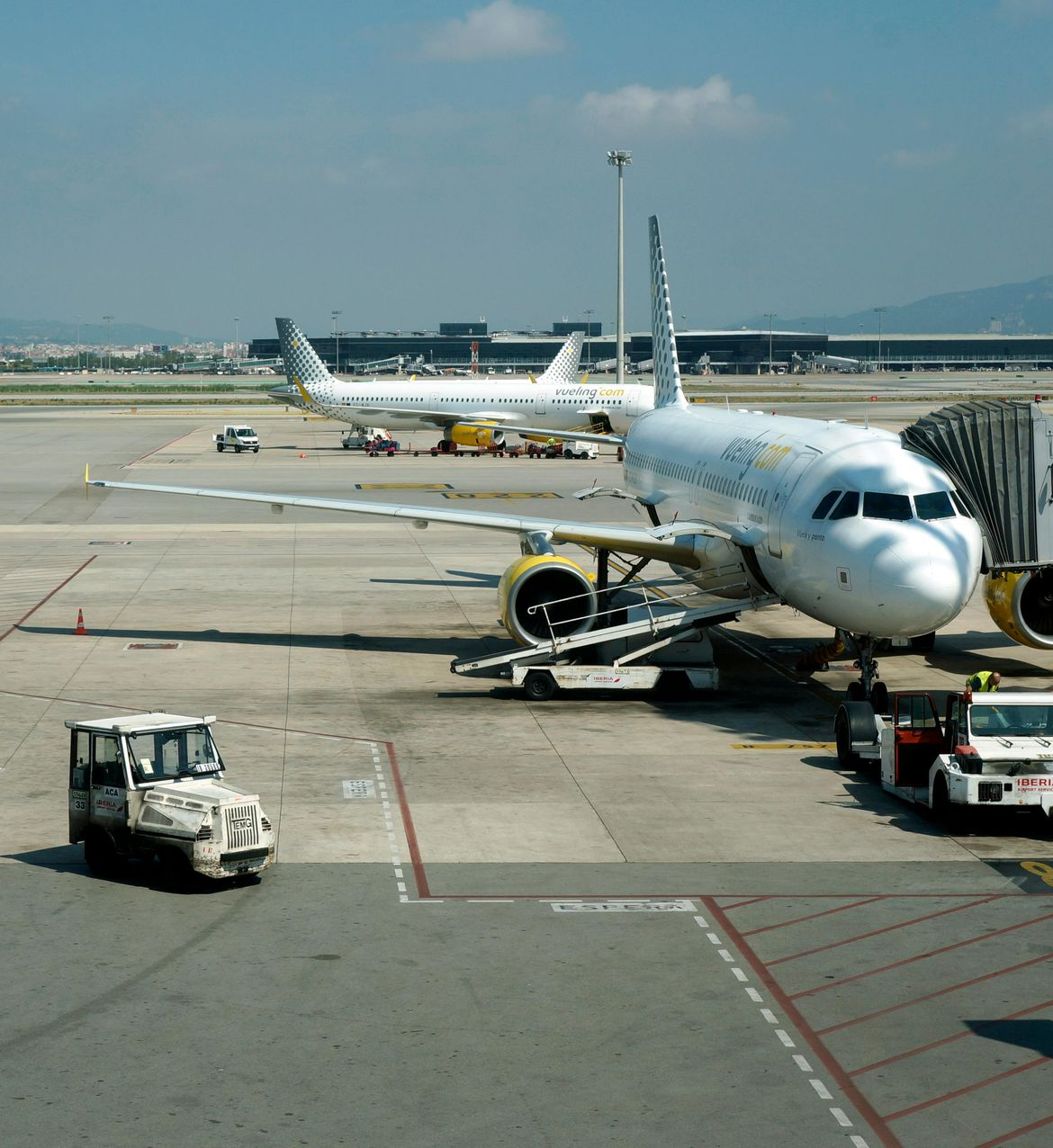 Airplane at airport gate with jet bridge, baggage carts, and another plane in the background