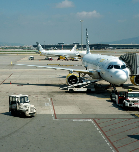 Airplane at airport gate with jet bridge, baggage carts, and another plane in the background