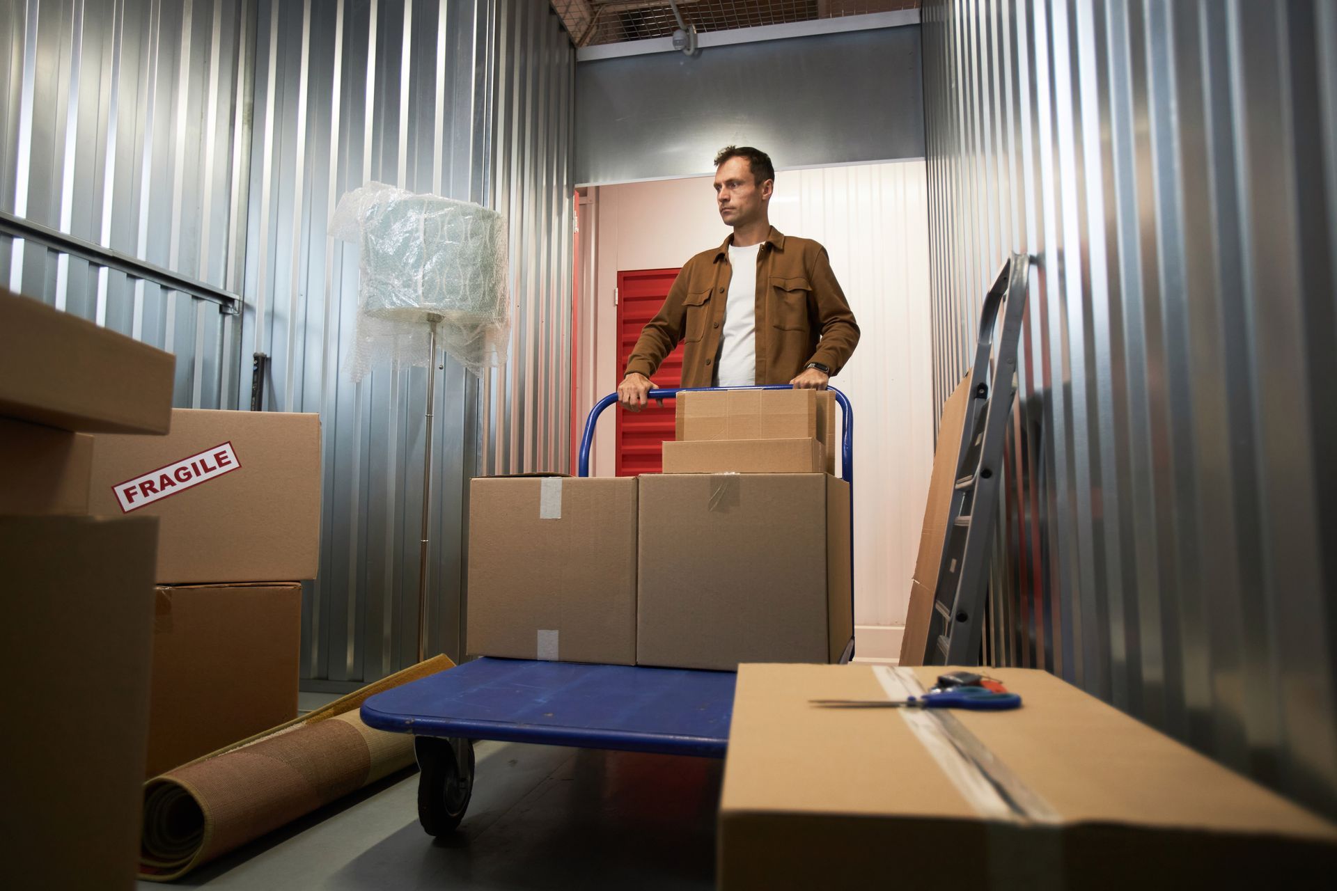 A pensive man pushing a cart with cardboard boxes inside a storage unit. A pensive man pushing a cart with cardboard boxes inside a storage unit.