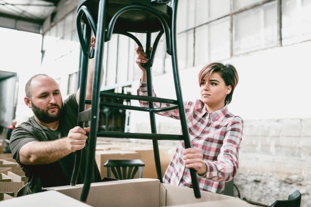 Two people handling a black chair in a warehouse with boxes and large industrial windows.
