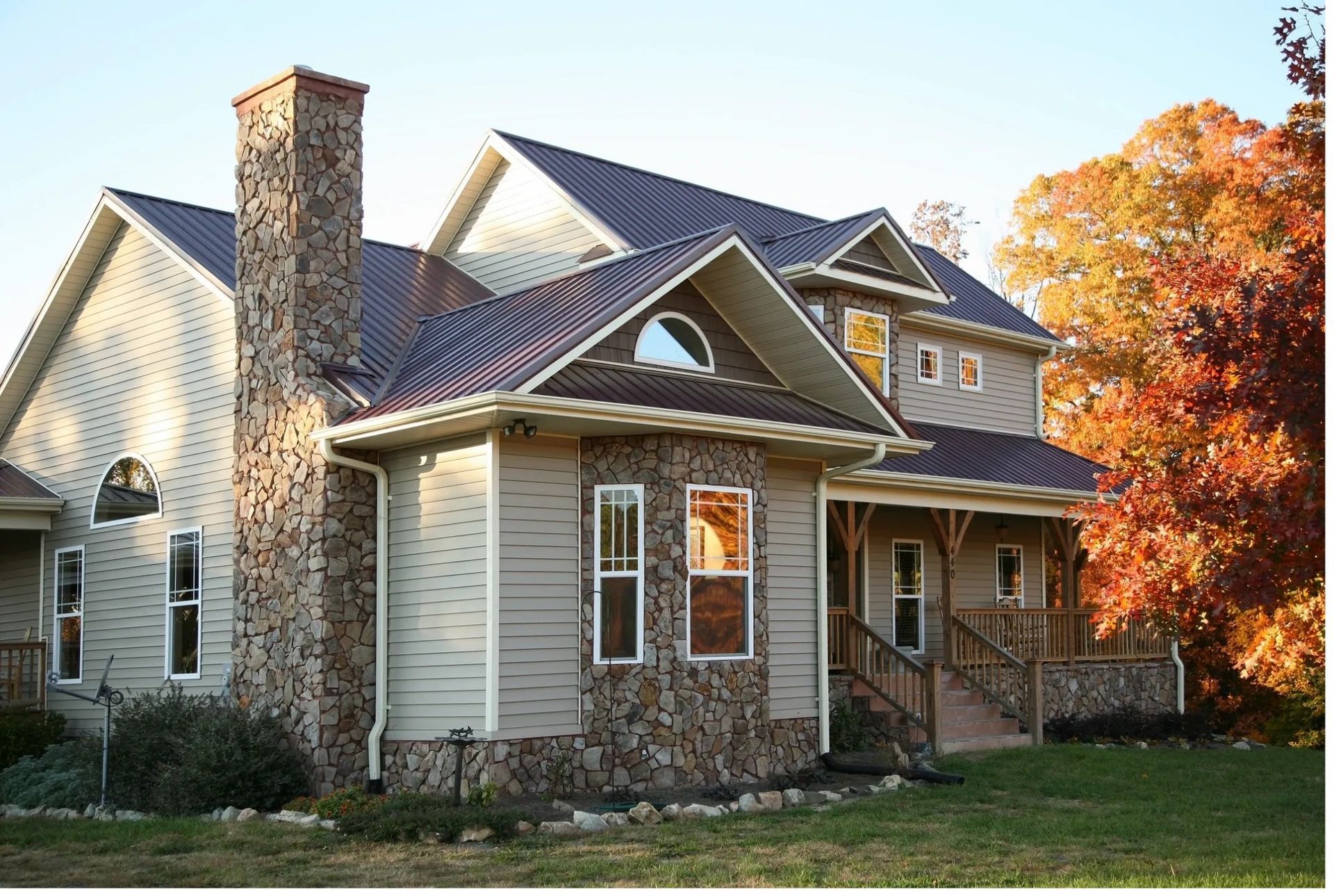Two-story house with stone chimney, tan siding, brown roof, and fall foliage in the yard.