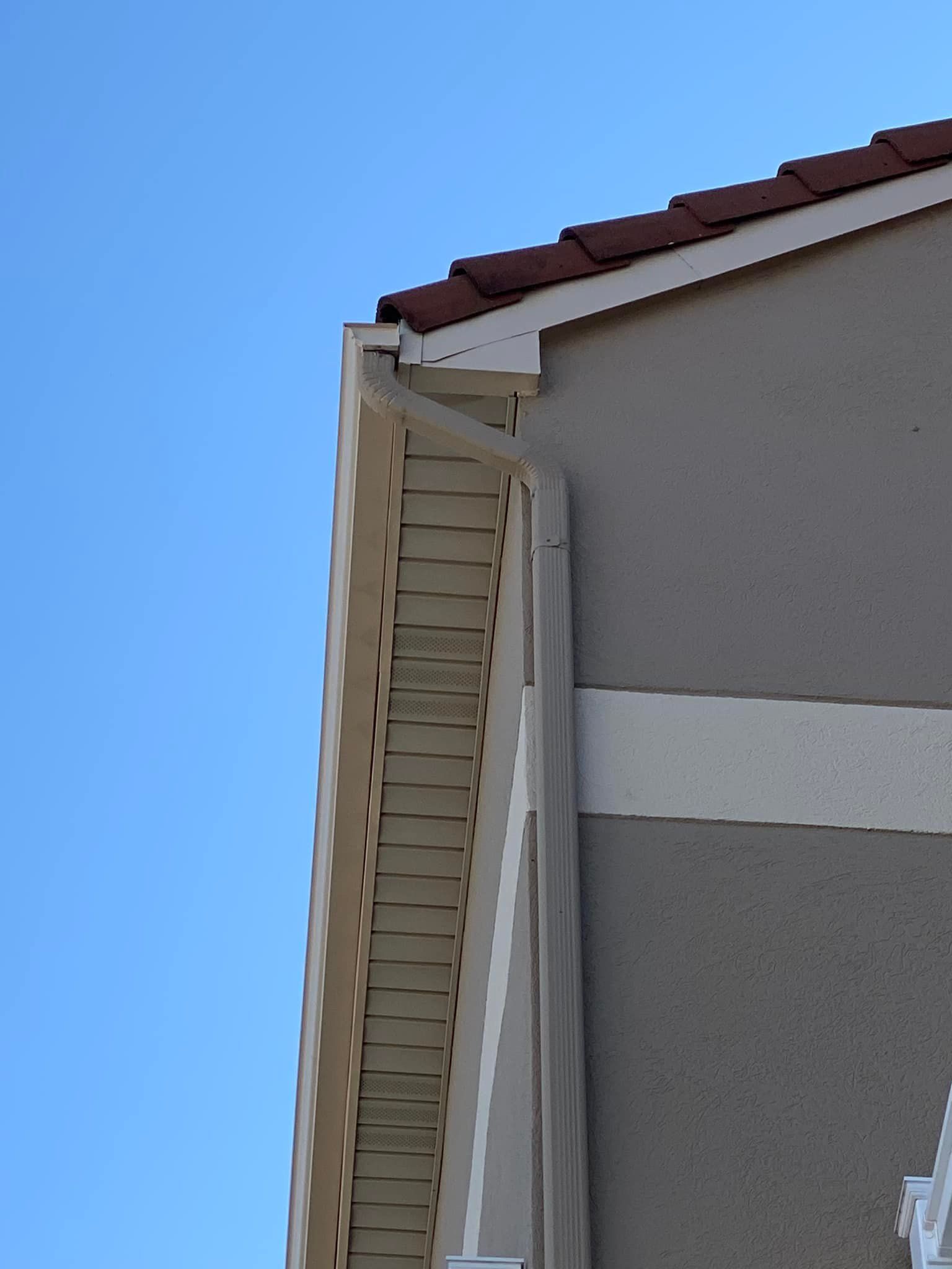 Corner of a tan stucco building with brown roof tiles and a light-colored trim against a blue sky.