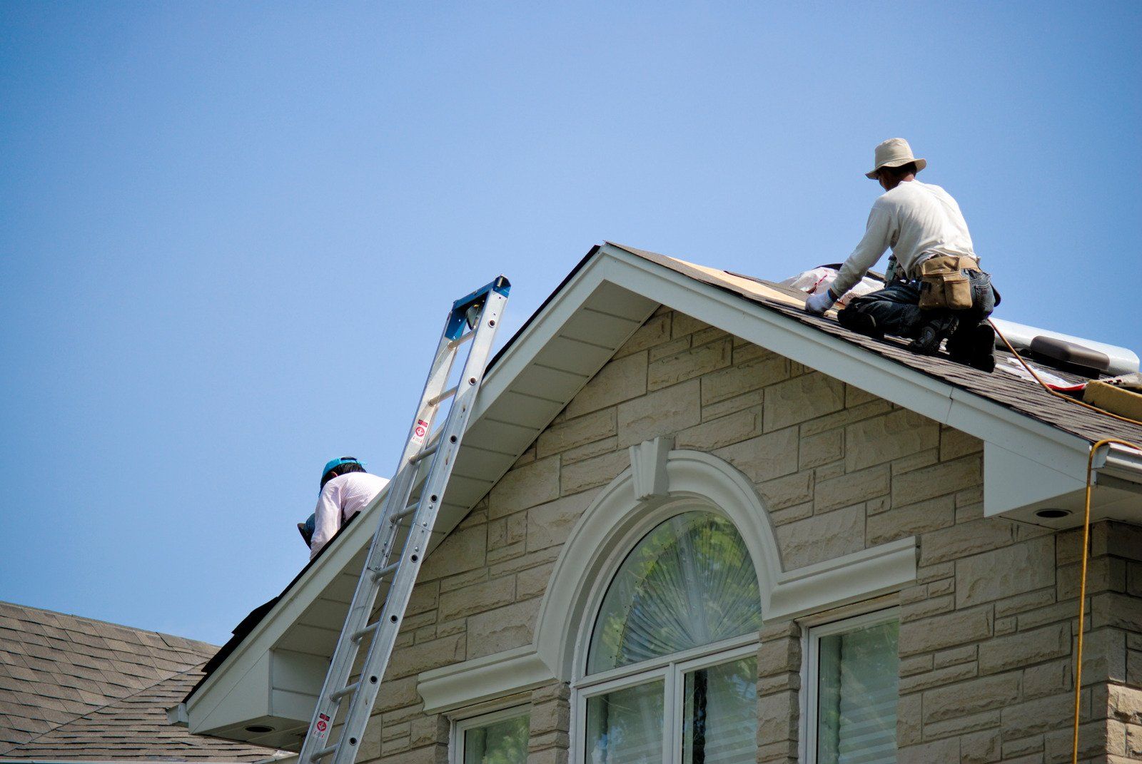Two roofers on a house roof under a blue sky; one on the roof, the other climbing a ladder.