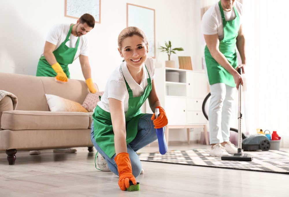 Three cleaners in green aprons, cleaning a living room. Woman smiles, kneeling to scrub floor. Men clean couch and vacuum.
