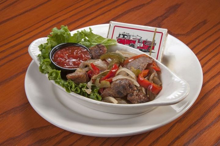 Sausage, peppers, and onions in a white dish with a side of tomato sauce and napkin, on a white plate.
