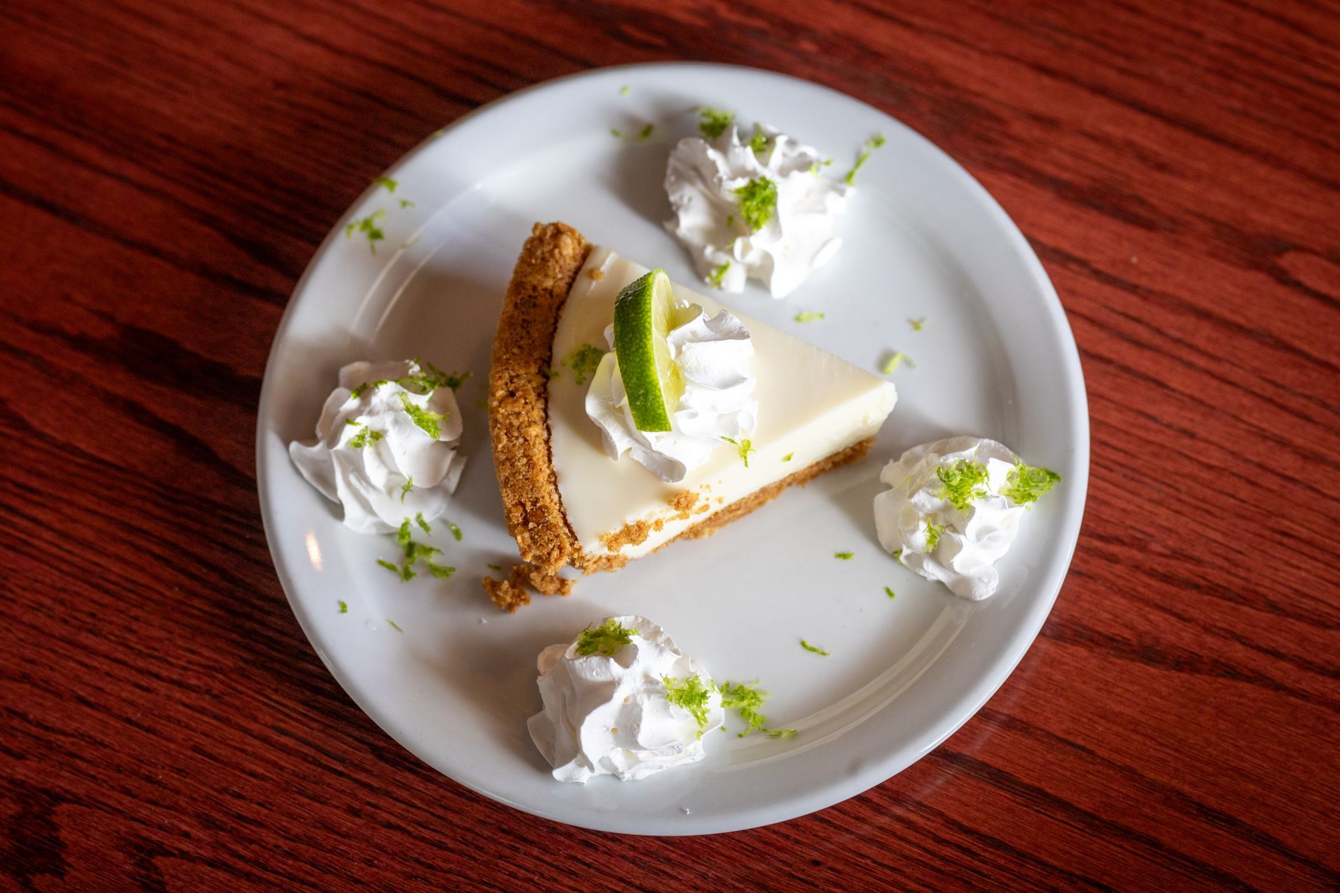 Slice of key lime pie on a white plate, topped with whipped cream and lime zest on a wooden table.