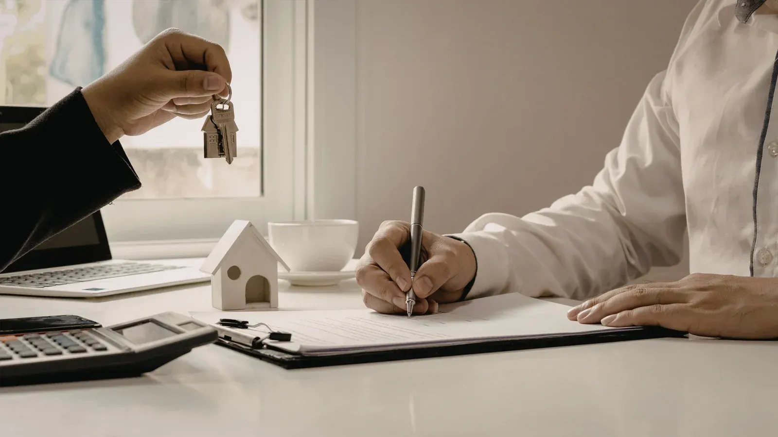Person handing keys as another signs a document, small house model on desk.