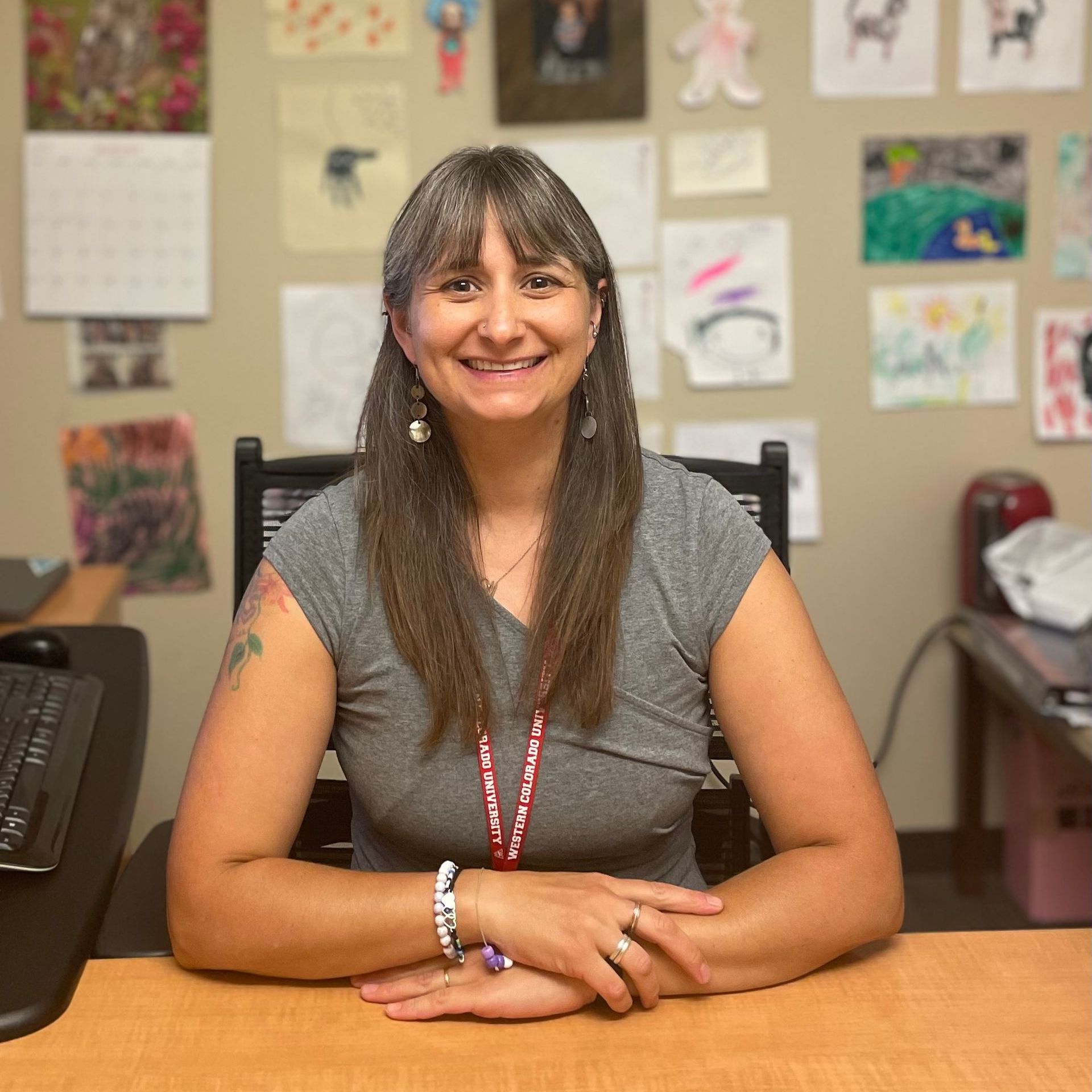 A woman is sitting at a desk with her hands folded and smiling.